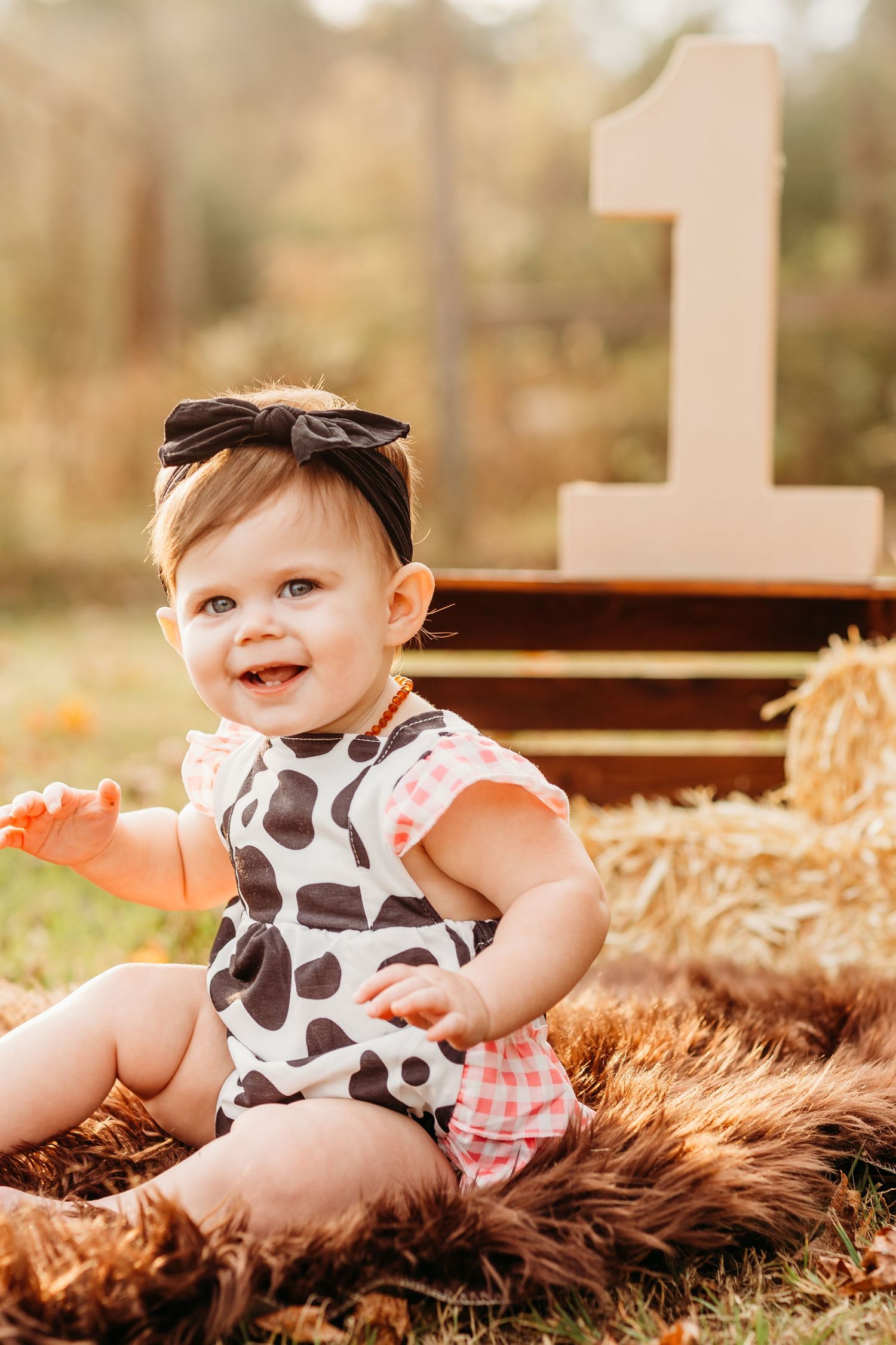 Baby in cow print outfit sits on furry rug with hay bales and wooden number one as backdrop for photo session.