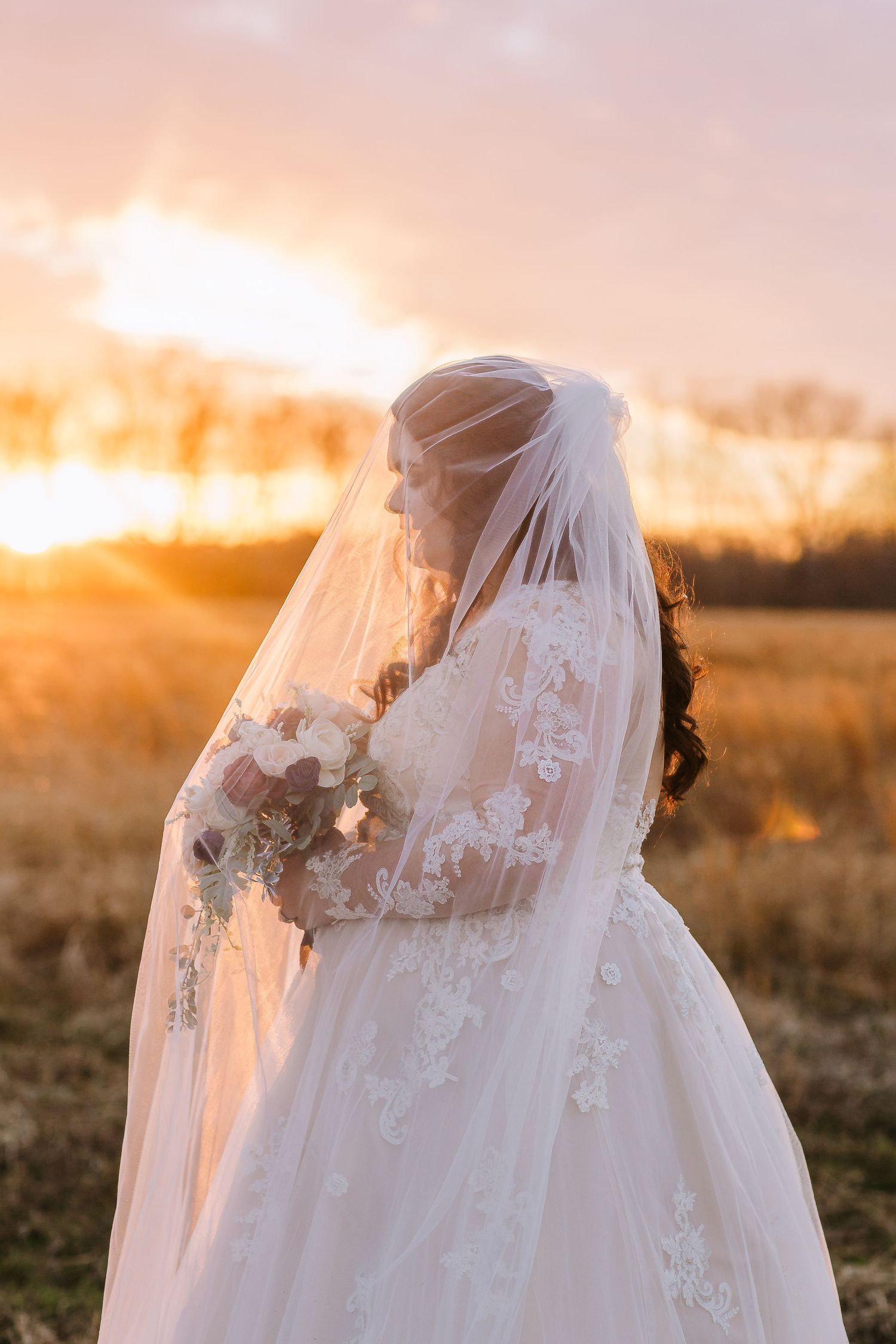 A bride in a lace wedding dress with long veil stands silhouetted against a golden sunset in a rural field.