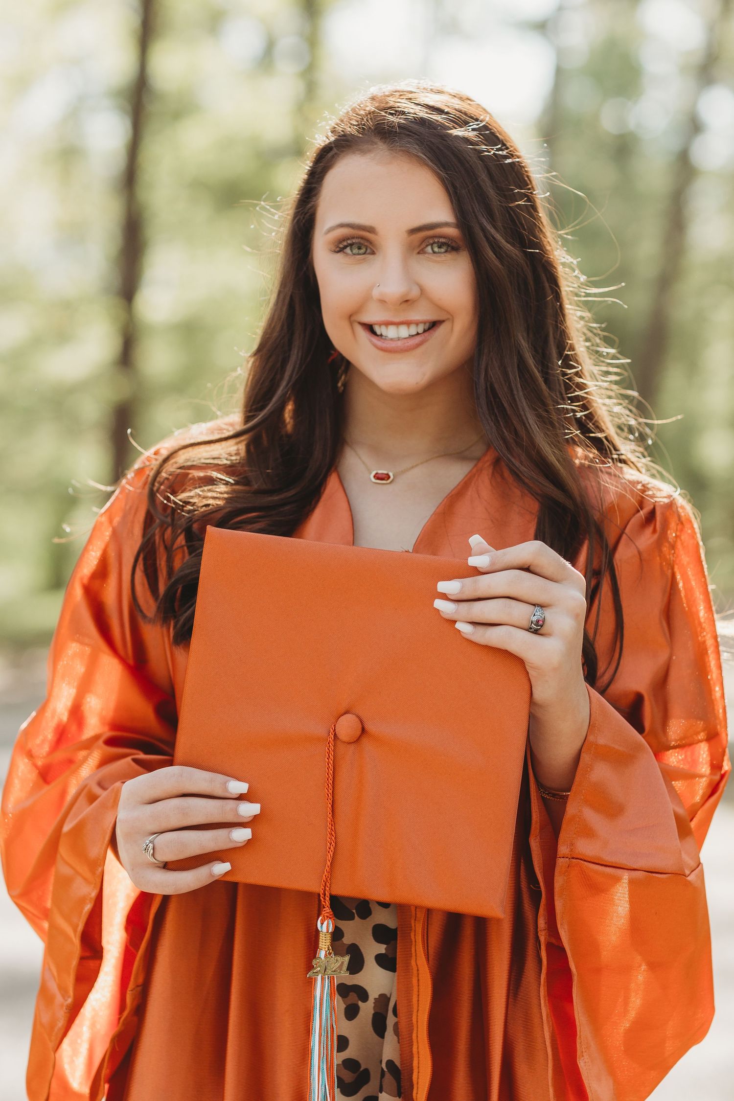 Graduate in bright orange academic gown holds up matching orange diploma holder outdoors on sunny day.
