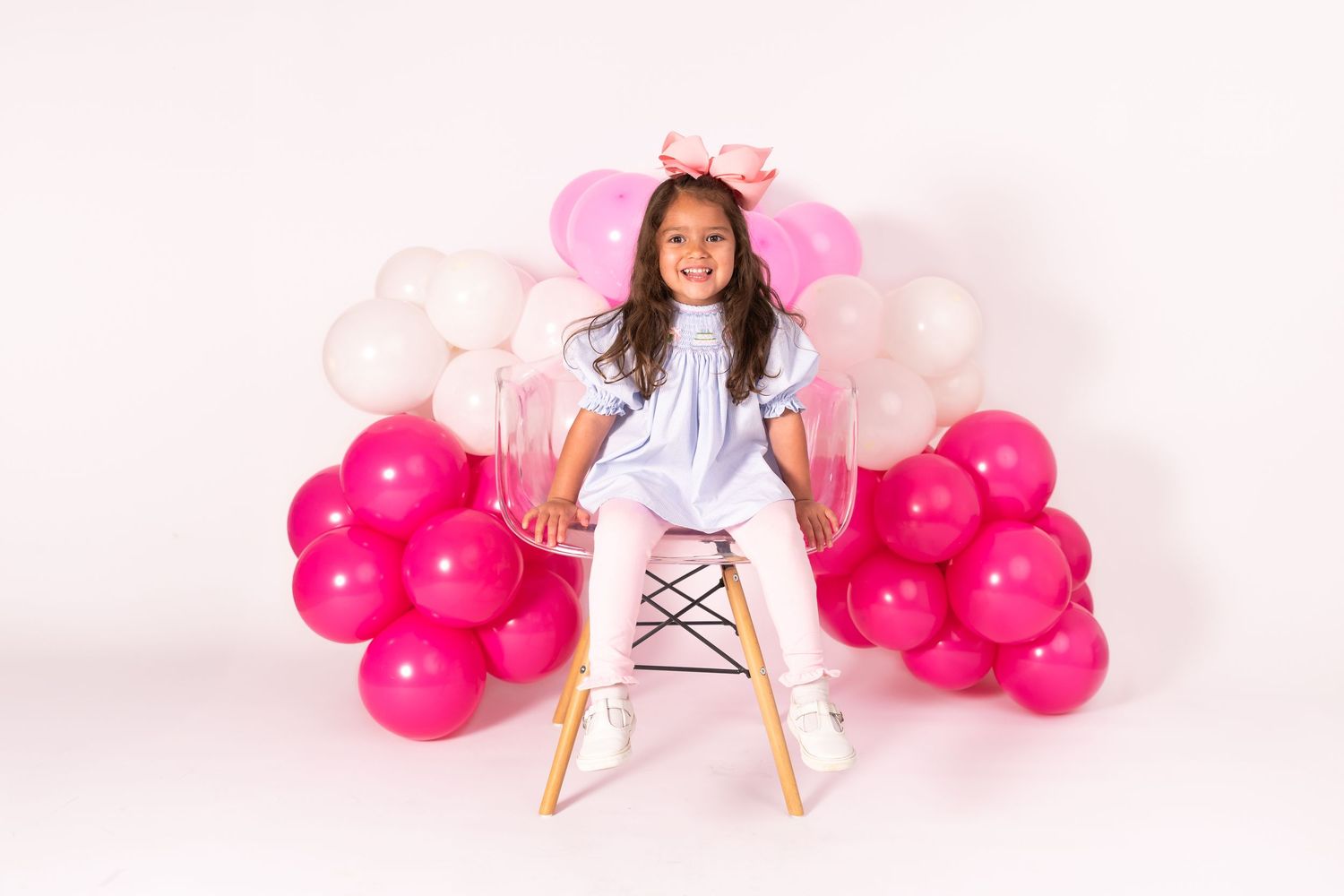 Sitting on high wooden chair with pink and white balloon arch display against white background.