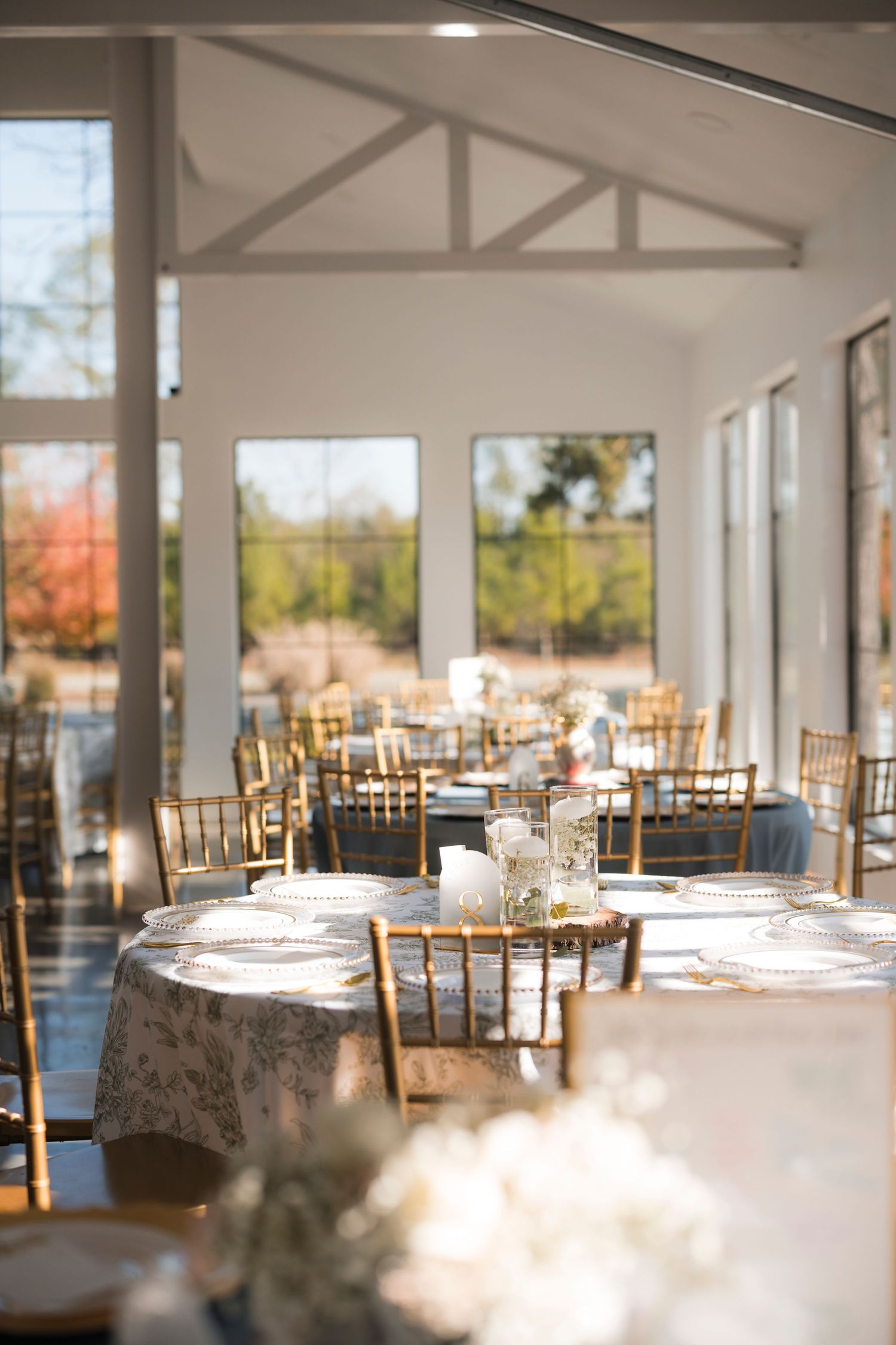 Elegant wedding reception hall with gold Chiavari chairs, floral tablecloths, and floor-to-ceiling windows overlooking autumn foliage.