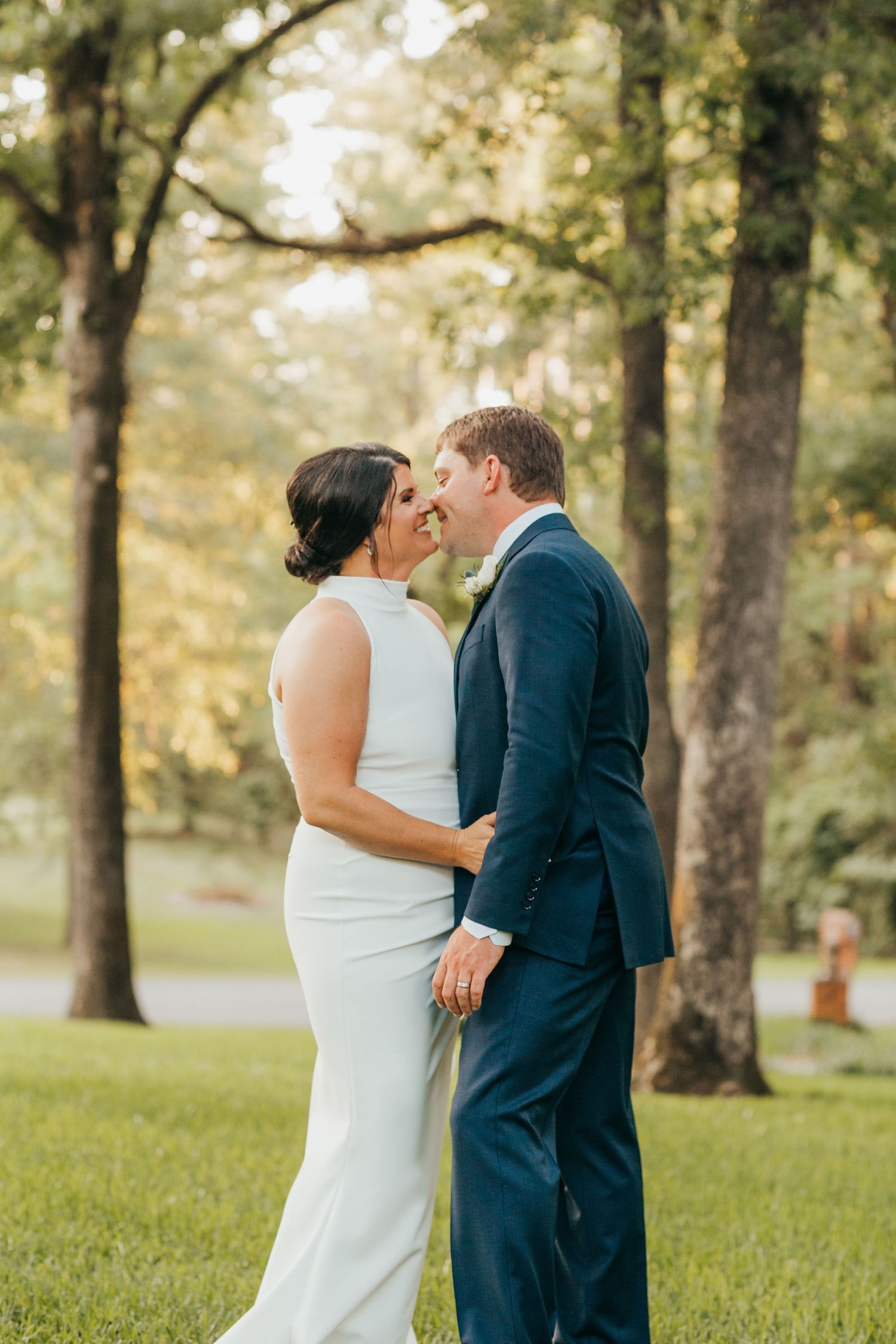A couple shares a romantic moment in a sun-dappled park setting with trees in the background.