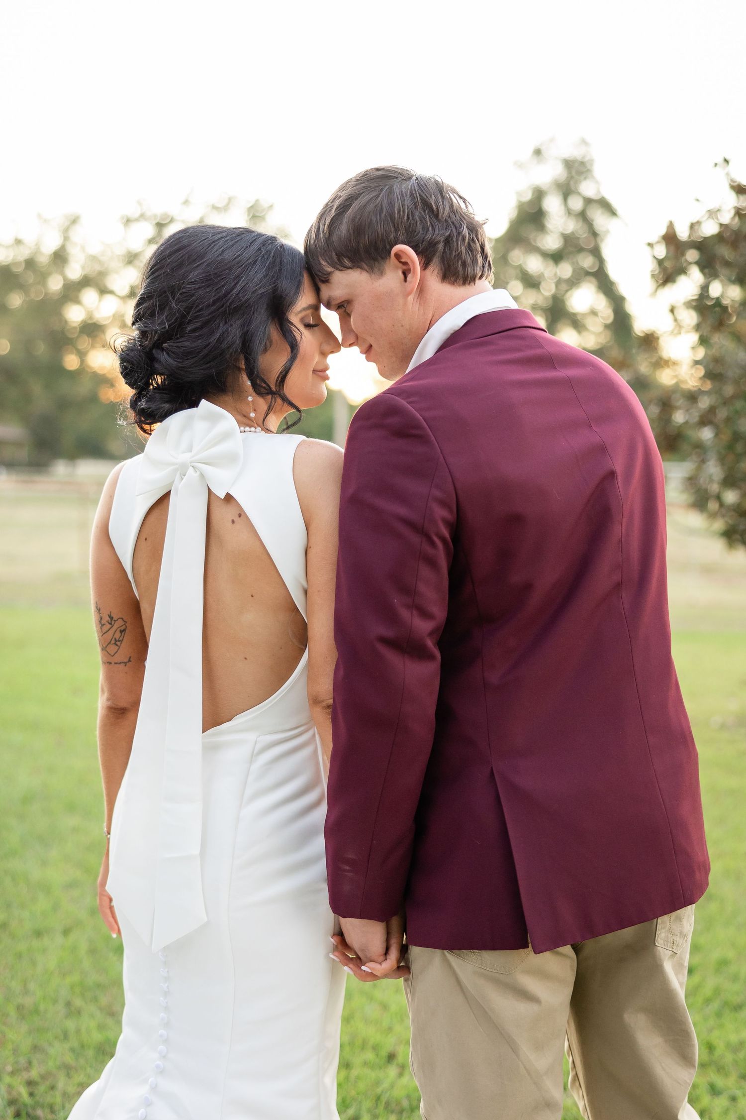 Bride in white open-back bow dress and groom in burgundy blazer hold hands, touching foreheads outdoors.