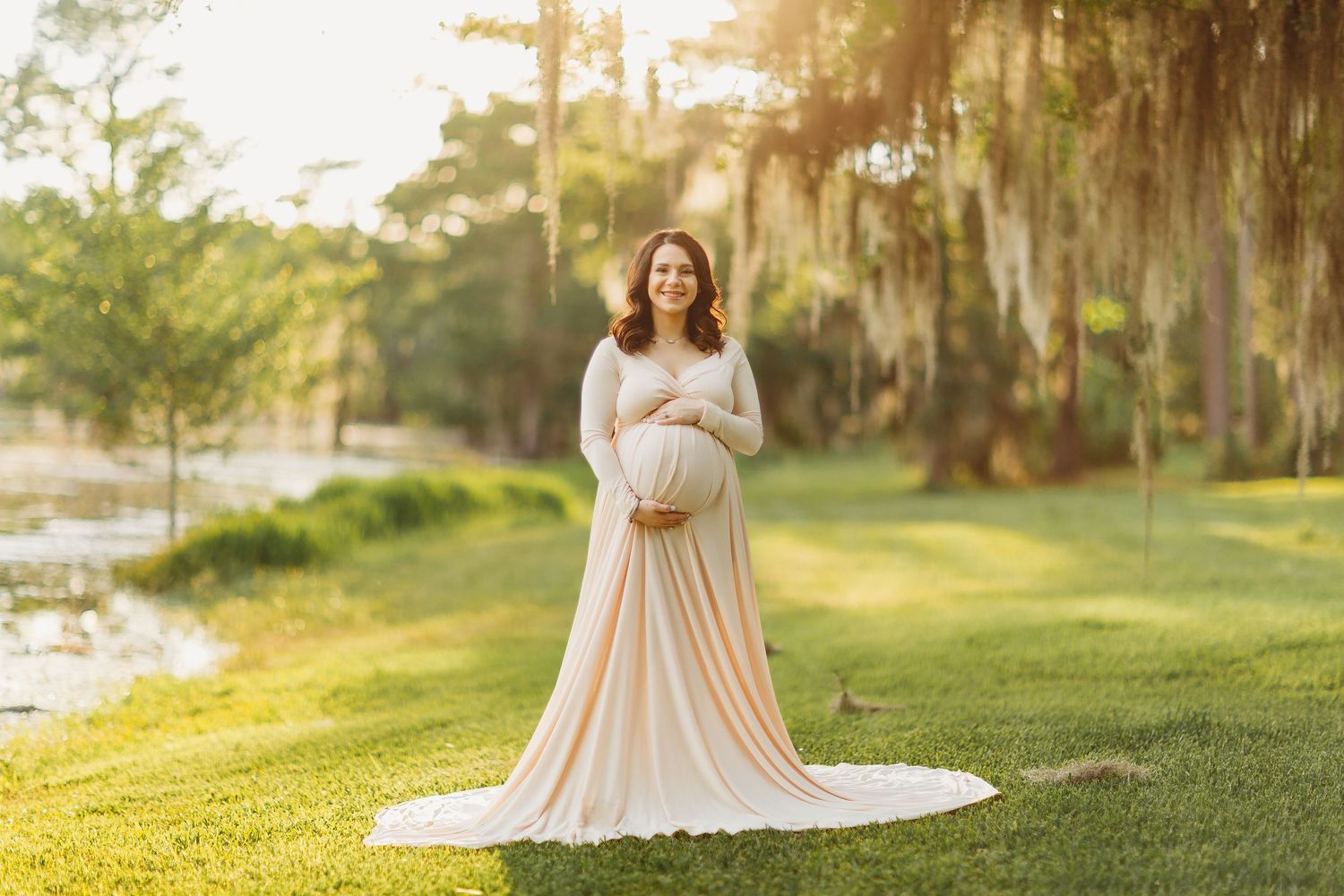 Expecting mother in flowing blush gown poses on grassy lawn at sunset with Spanish moss draped trees in background.