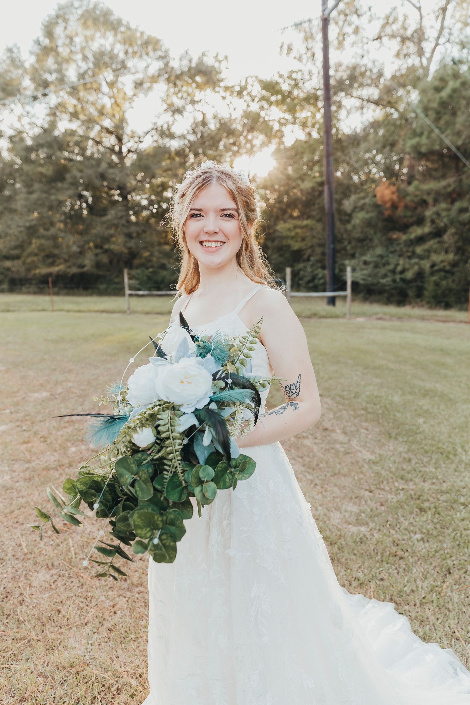 A bride in a flowing white gown holds a lush bouquet of eucalyptus and white flowers in an outdoor garden setting at sunset.