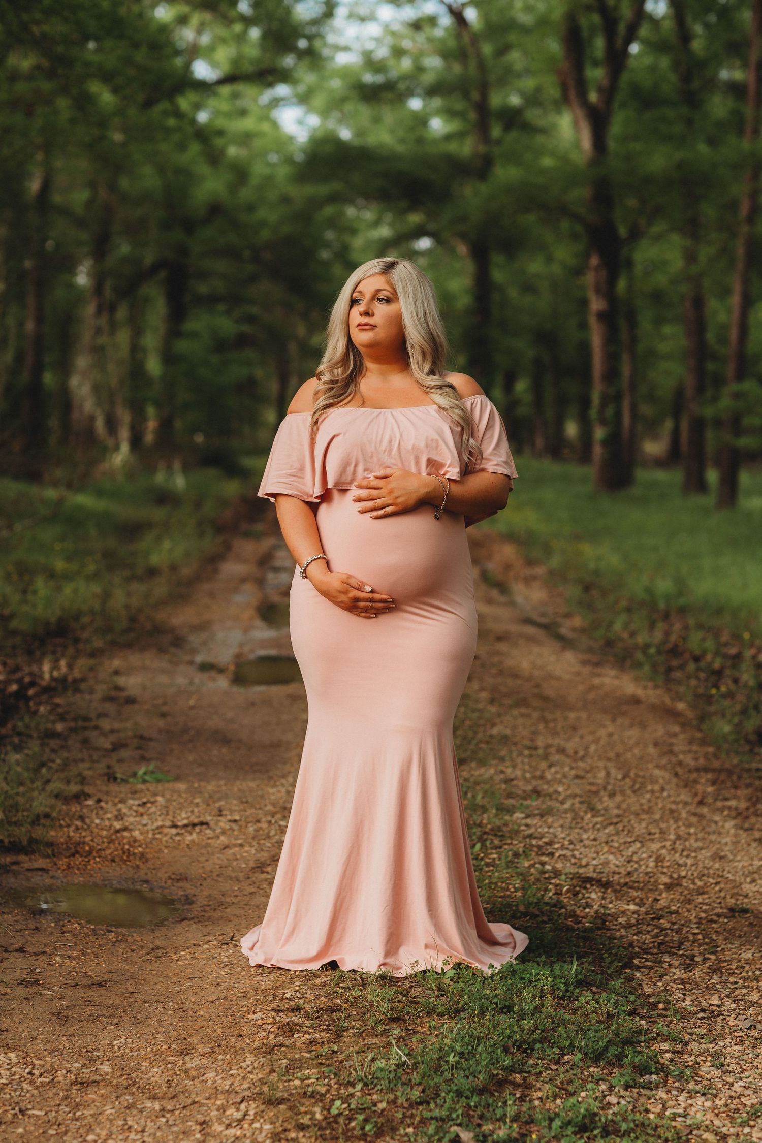 An expectant woman in a flowing blush pink dress poses on a wooded dirt path during a maternity photoshoot.