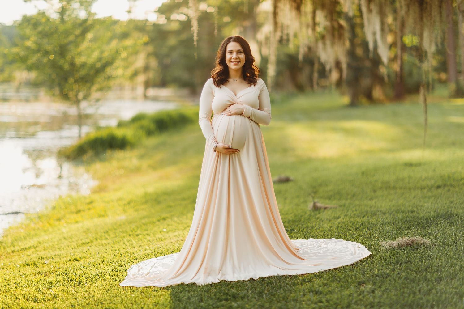Maternity photo of an elegant cream-colored flowing dress worn outdoors by a lake with Spanish moss and golden sunlight.