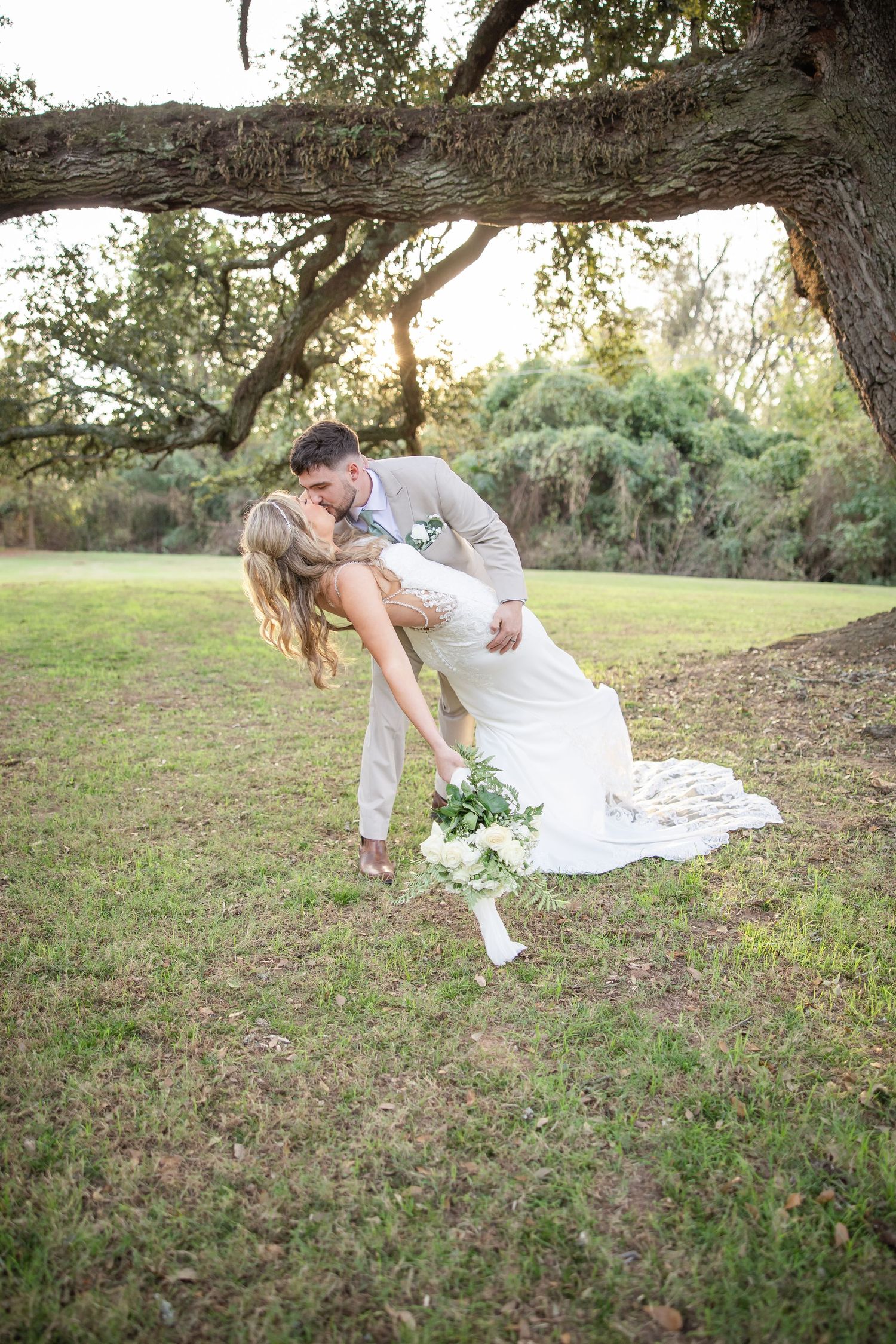 Bride and groom share a romantic dip kiss under a sprawling oak tree during golden hour outdoor wedding.