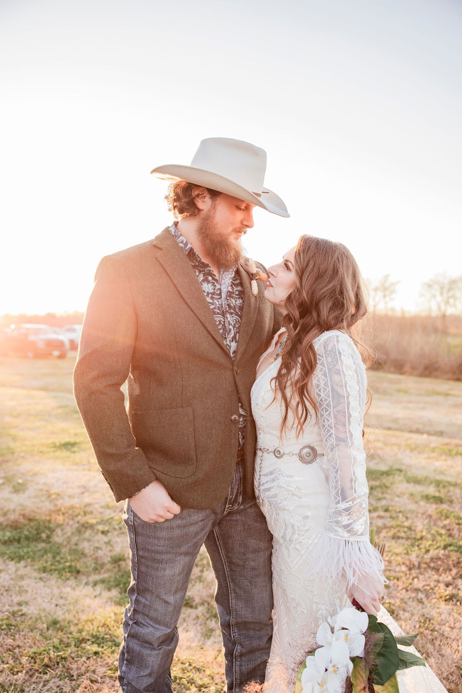 A romantic western-style couple embraces in a golden sunset field wearing cowboy attire and wedding dress.