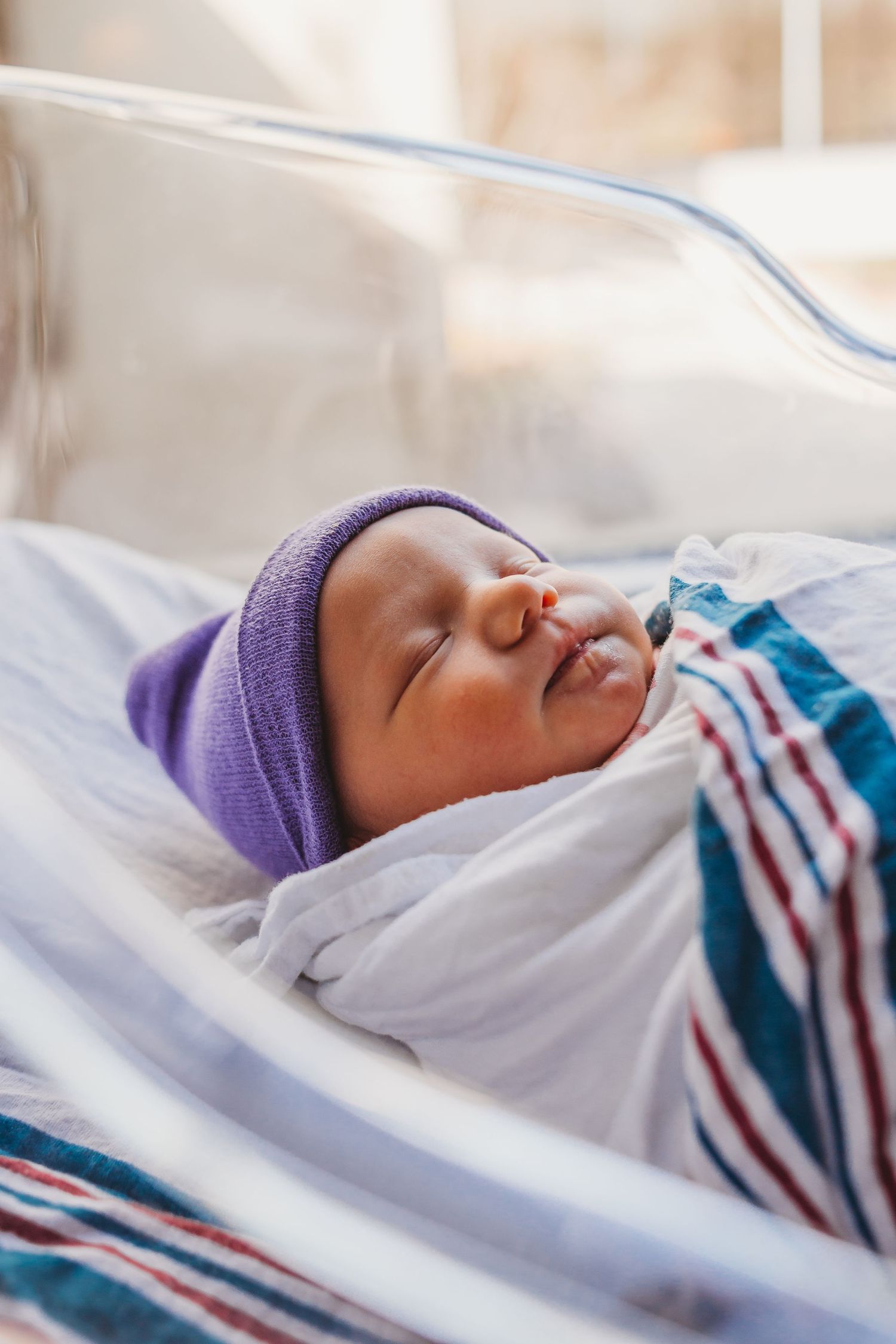 Peaceful sleeping newborn wearing purple cap and striped blanket rests in hospital bassinet.