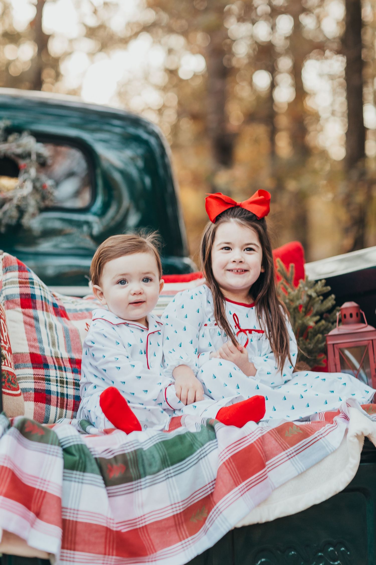 Two children in Christmas pajamas sit in the back of a vintage car with plaid blankets and holiday decorations.