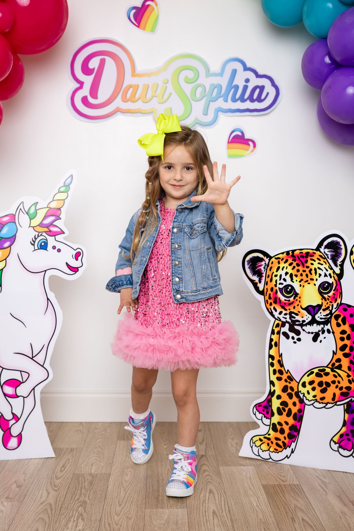 Pink party setting with colorful balloon arch, unicorn and leopard decorations against white wall.