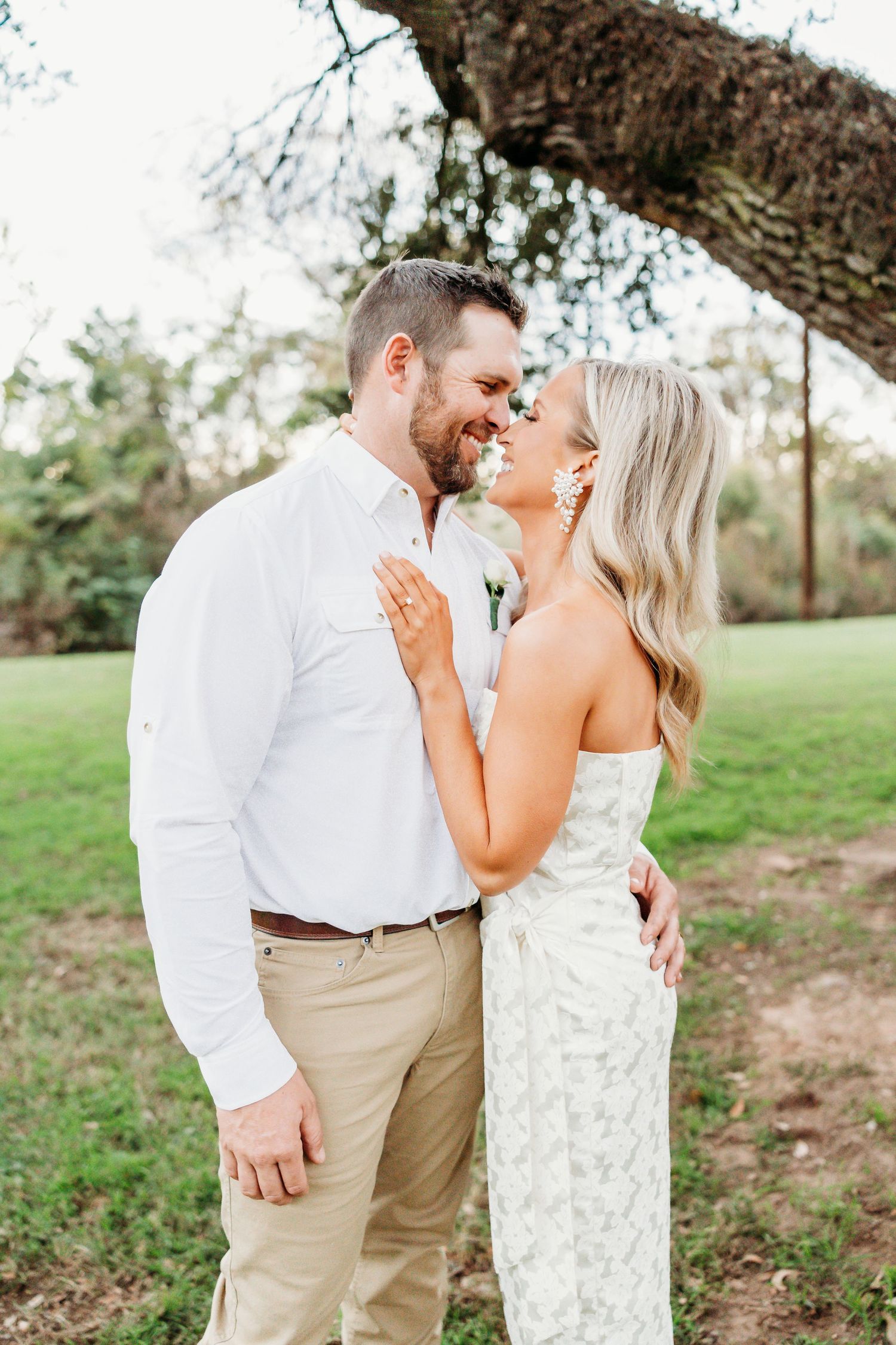 A smiling couple in white outfits share a romantic moment outdoors under a tree on their wedding day.