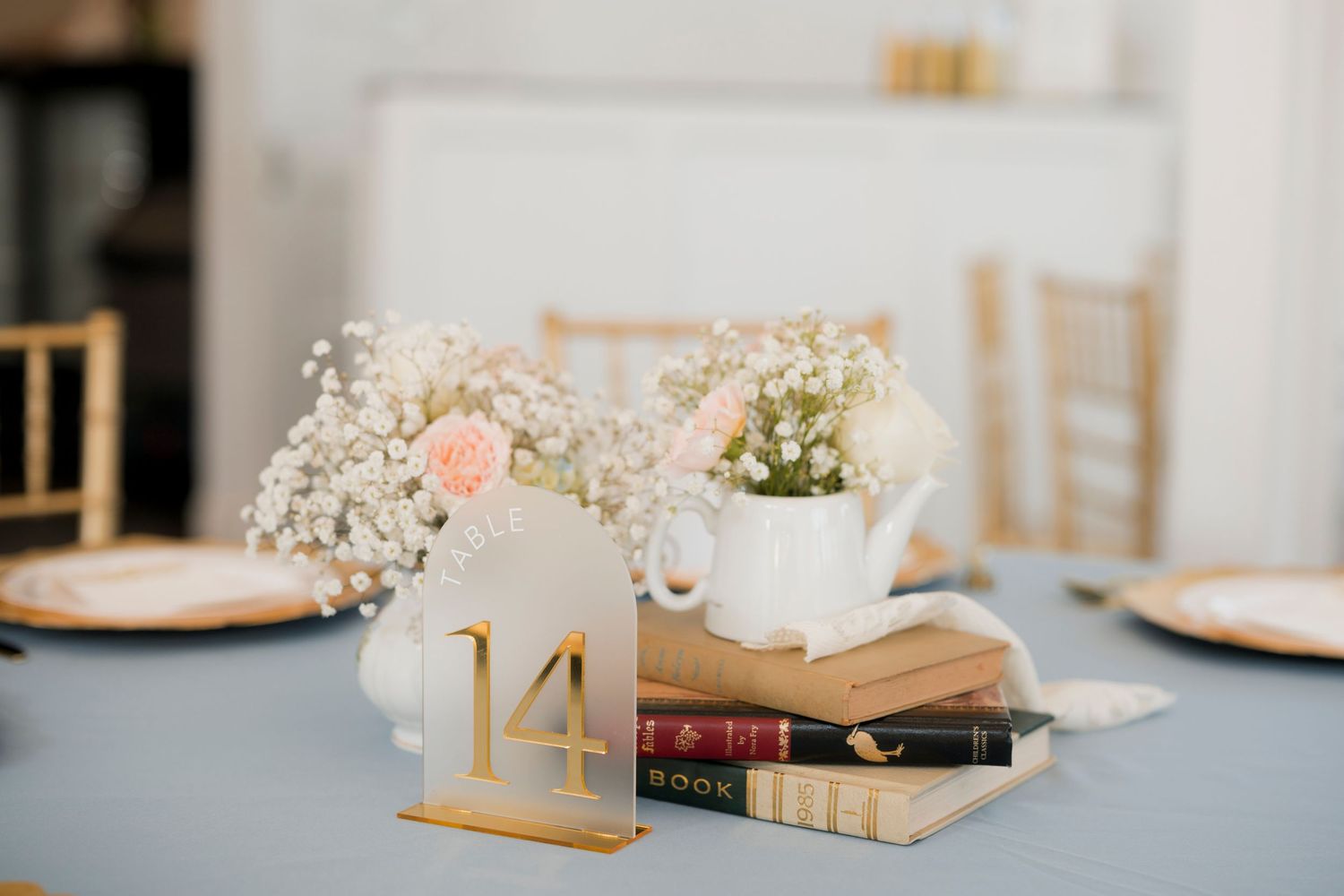 Wedding table centerpiece with baby's breath, pink roses in a white watering can, stacked books, and gold number 14 sign.
