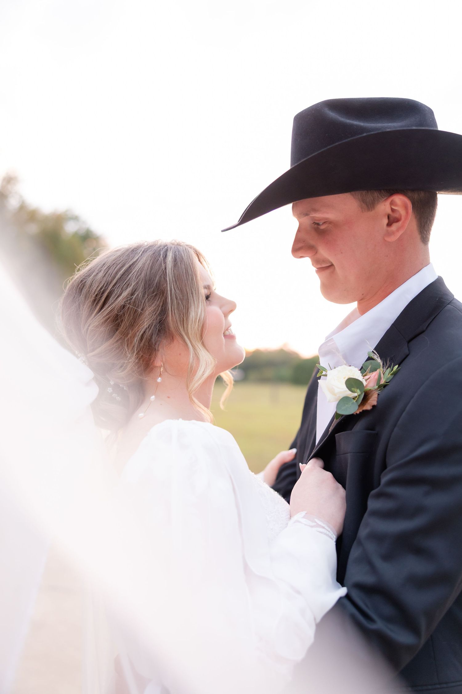 A bride and groom share a romantic moment outdoors, with the groom wearing a black cowboy hat and boutonniere.