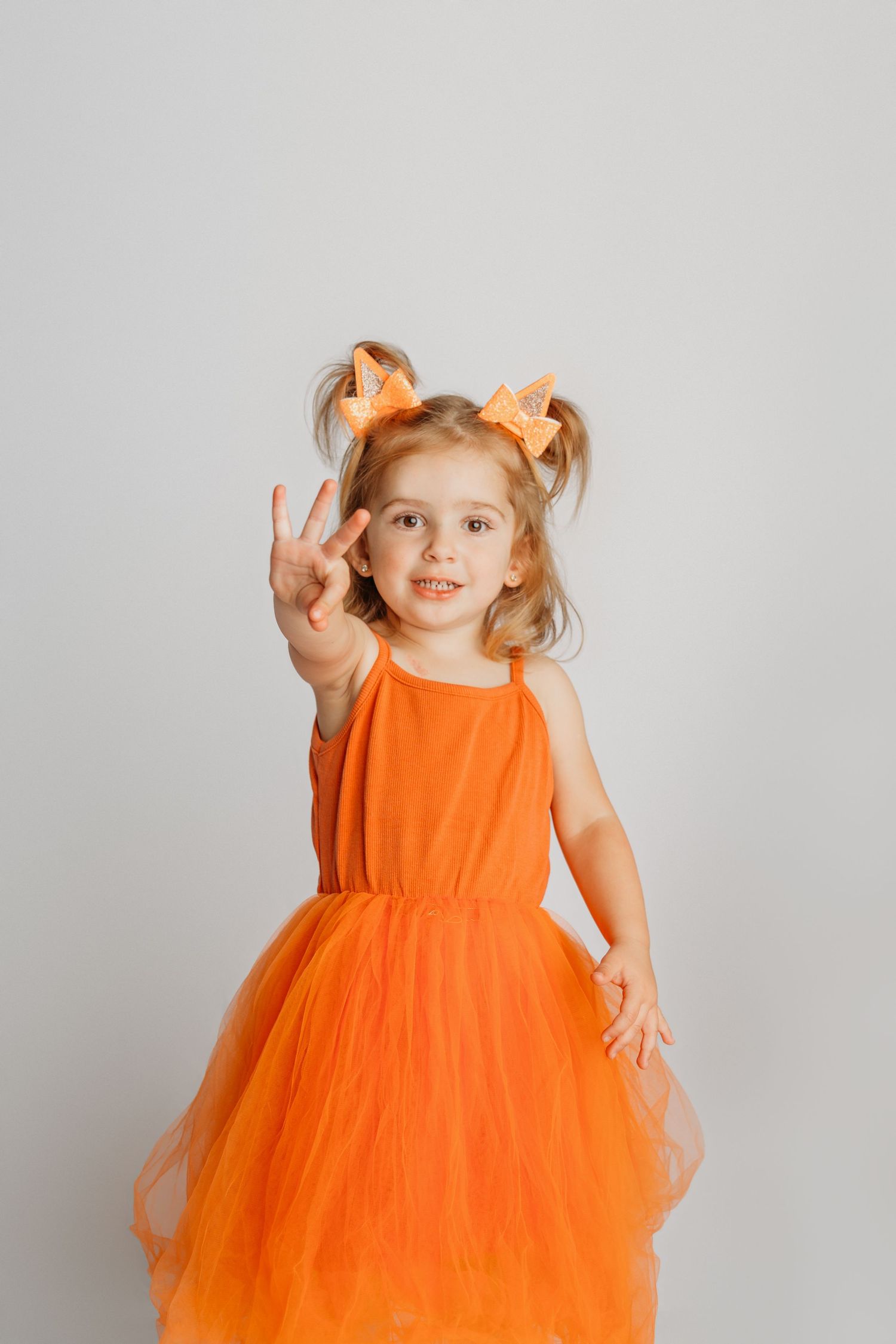 Child in vibrant orange tulle dress posing playfully against a plain grey background.
