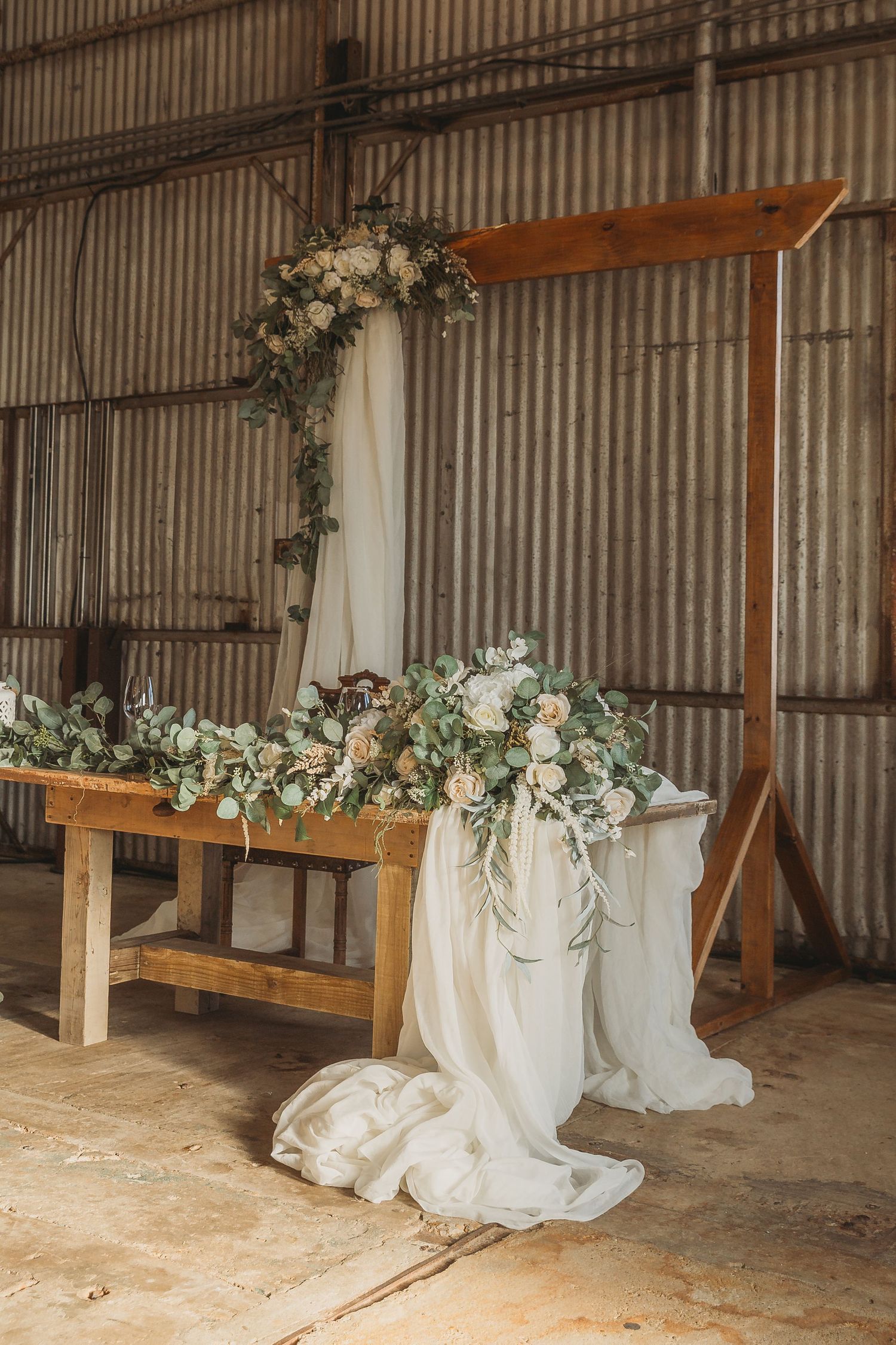 Rustic wooden wedding arch decorated with flowing white fabric and elegant floral arrangements in an industrial venue.