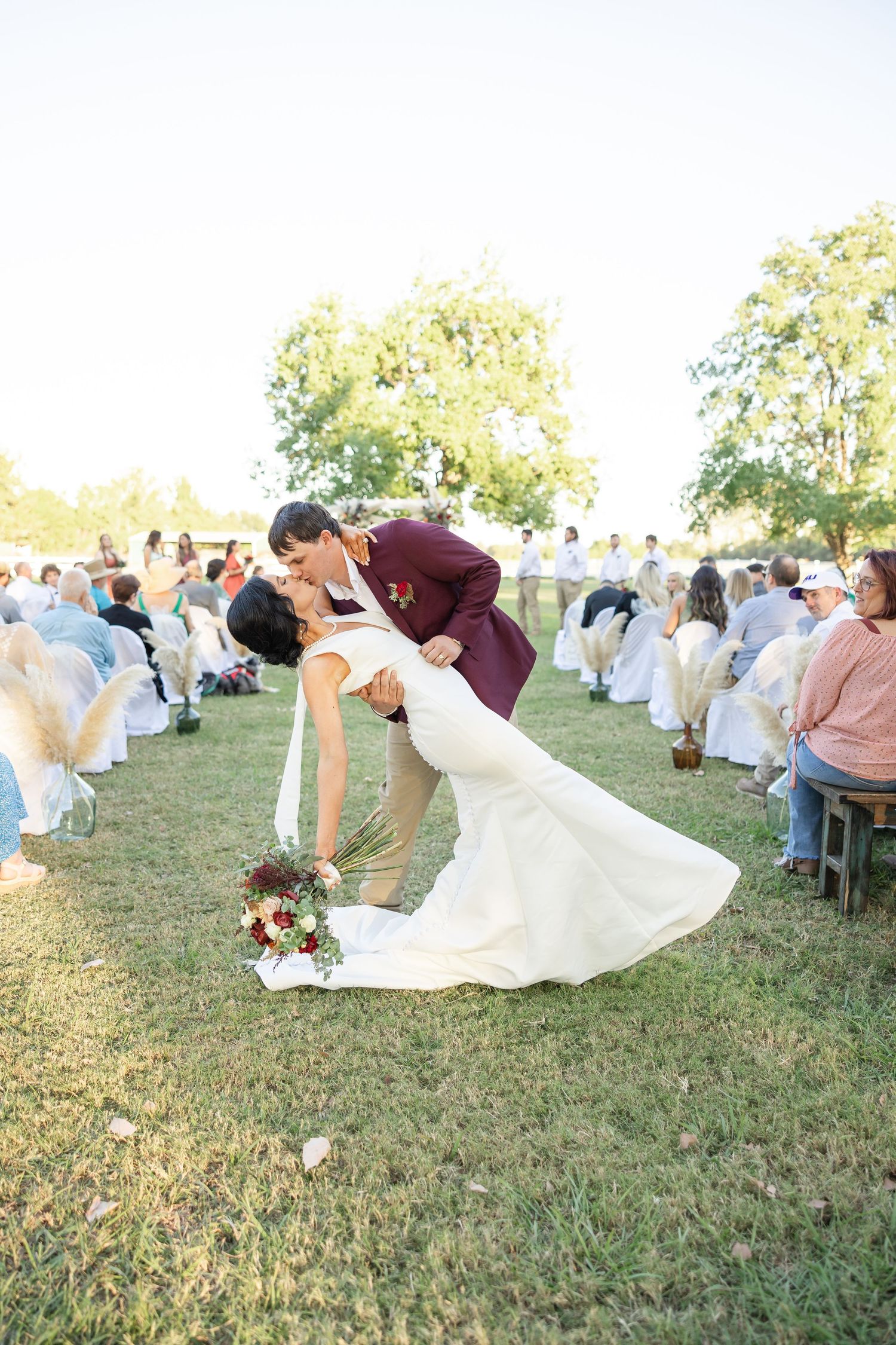 Groom in burgundy suit dips bride in white gown for a kiss at outdoor wedding ceremony on green lawn.