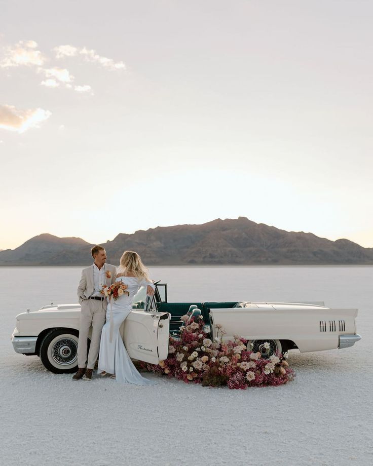 A couple stands beside a classic white convertible car decorated with flowers on a salt flat with mountains in the background.