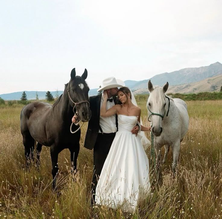 A couple in wedding attire poses with two horses in a scenic mountain meadow with tall grass and distant peaks.