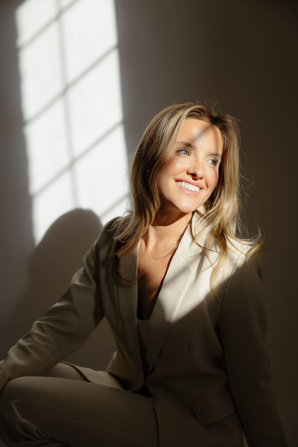 Professional fashion portrait with dramatic lighting and shadows showing a model in a dark blazer against a white wall.