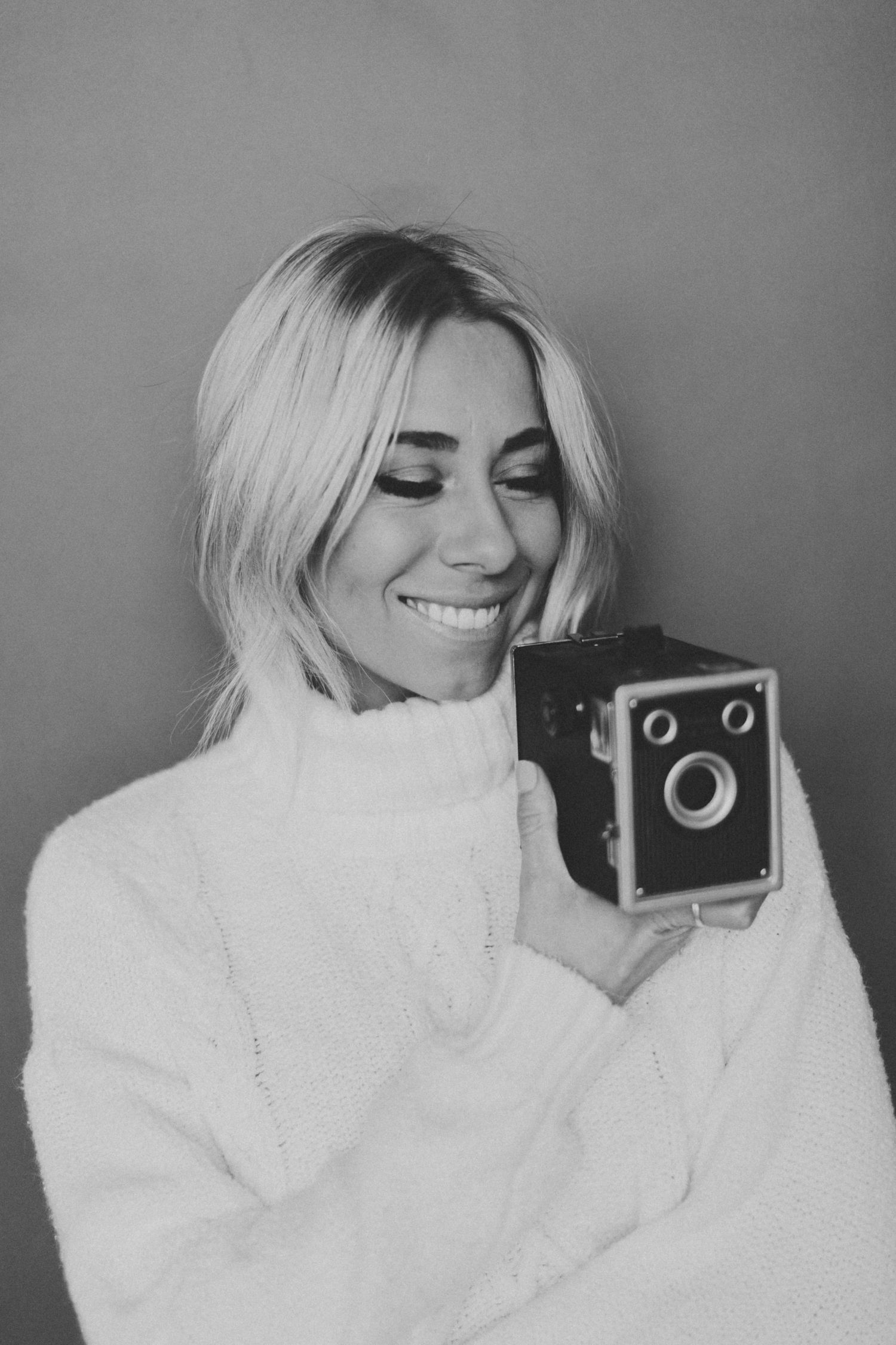 A series of black and white photos showing someone in a white sweater posing with a vintage camera against a gray background.