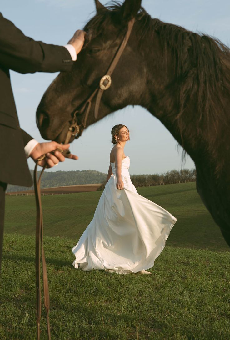 A white wedding dress flows in the breeze as a dark horse stands beside a person in a grassy field at sunset.