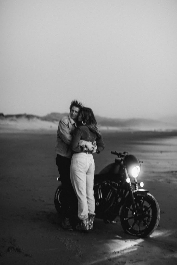 A romantic black and white photo of a couple embracing next to a motorcycle on a misty beach at dusk.