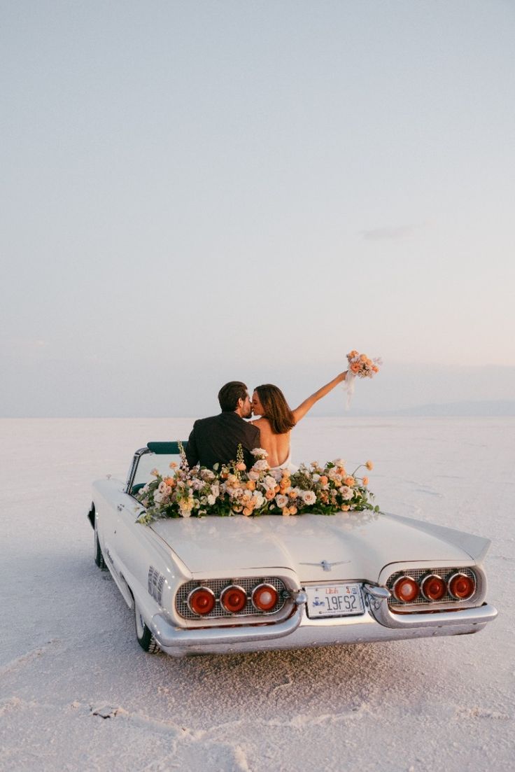 Vintage white convertible car parked on white salt flats with a floral arrangement and couple at sunset.