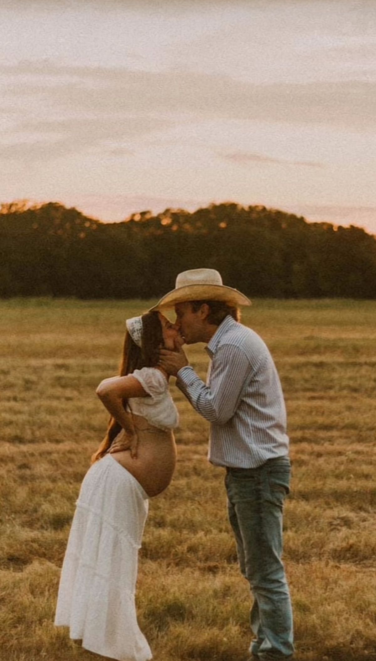 A couple sharing a romantic kiss in a field at sunset with the man wearing a cowboy hat.