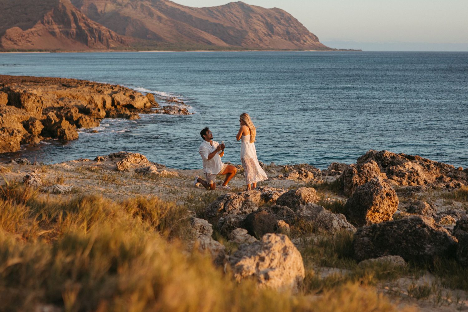 Man proposing to woman on rocky coastline at golden hour with mountains and turquoise sea in background.