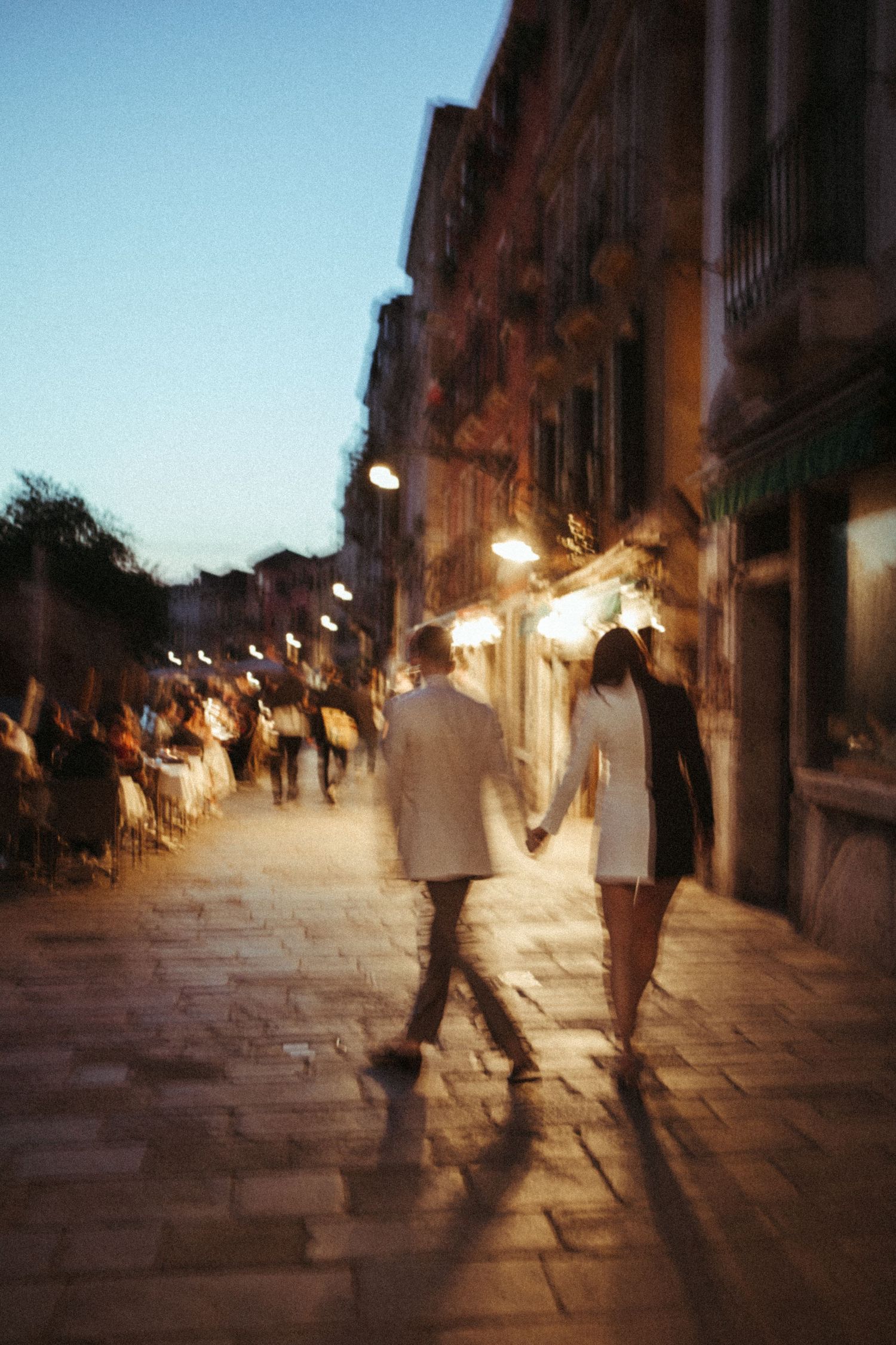 Couple holding hands walking cobblestone street at dusk in Venice, Italy.