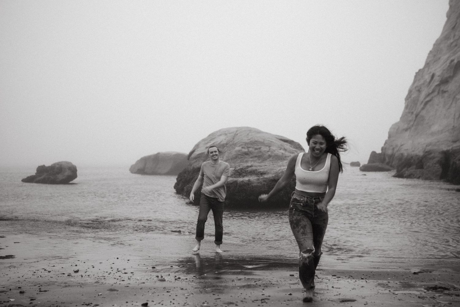 Two women running on a foggy beach with large rocks and misty ocean in a black and white photo.