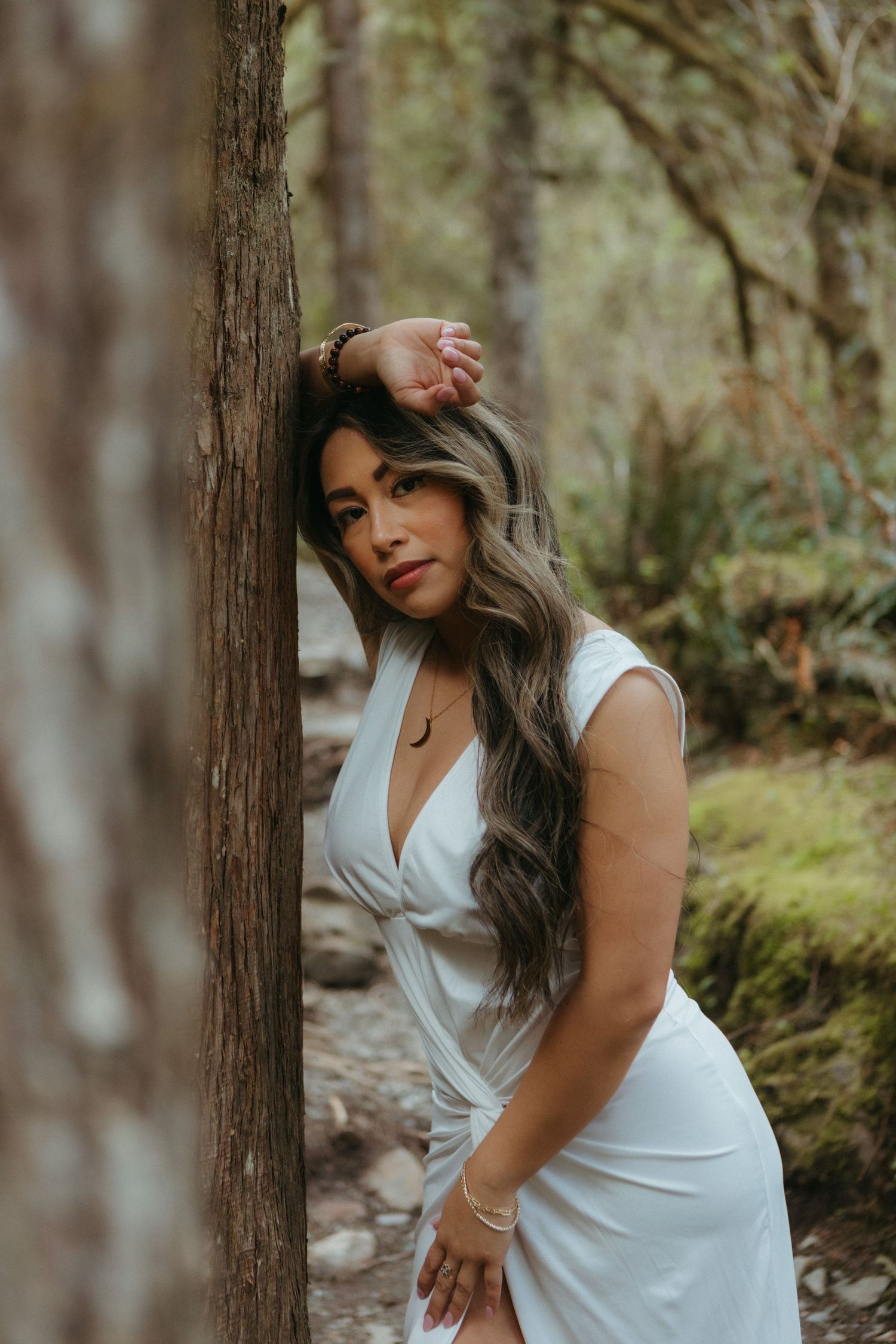 A person in a white dress poses gracefully against tree trunks in a misty forest setting.