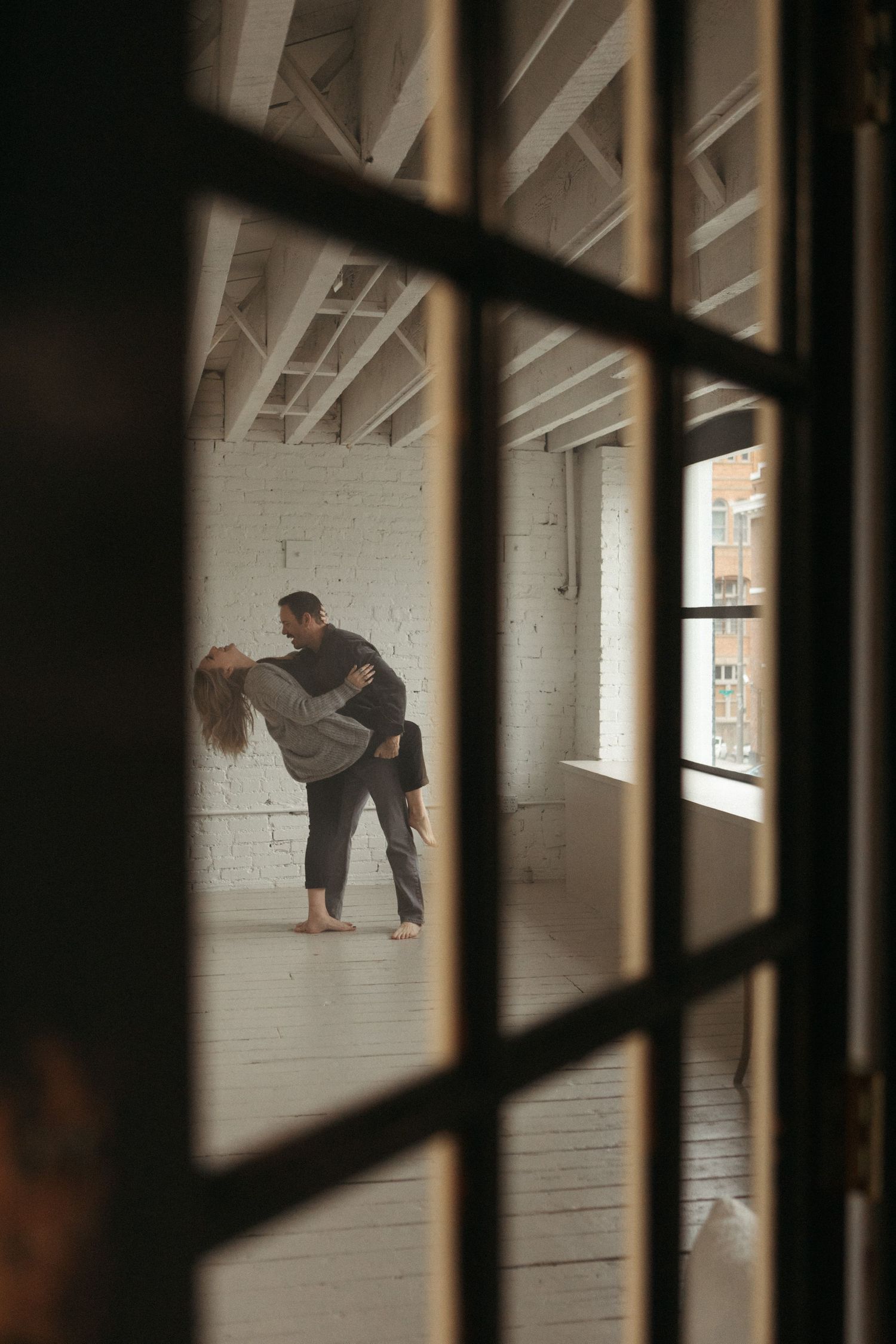 A couple dancing romantically in a bright loft studio, viewed through a glass door with black frames.