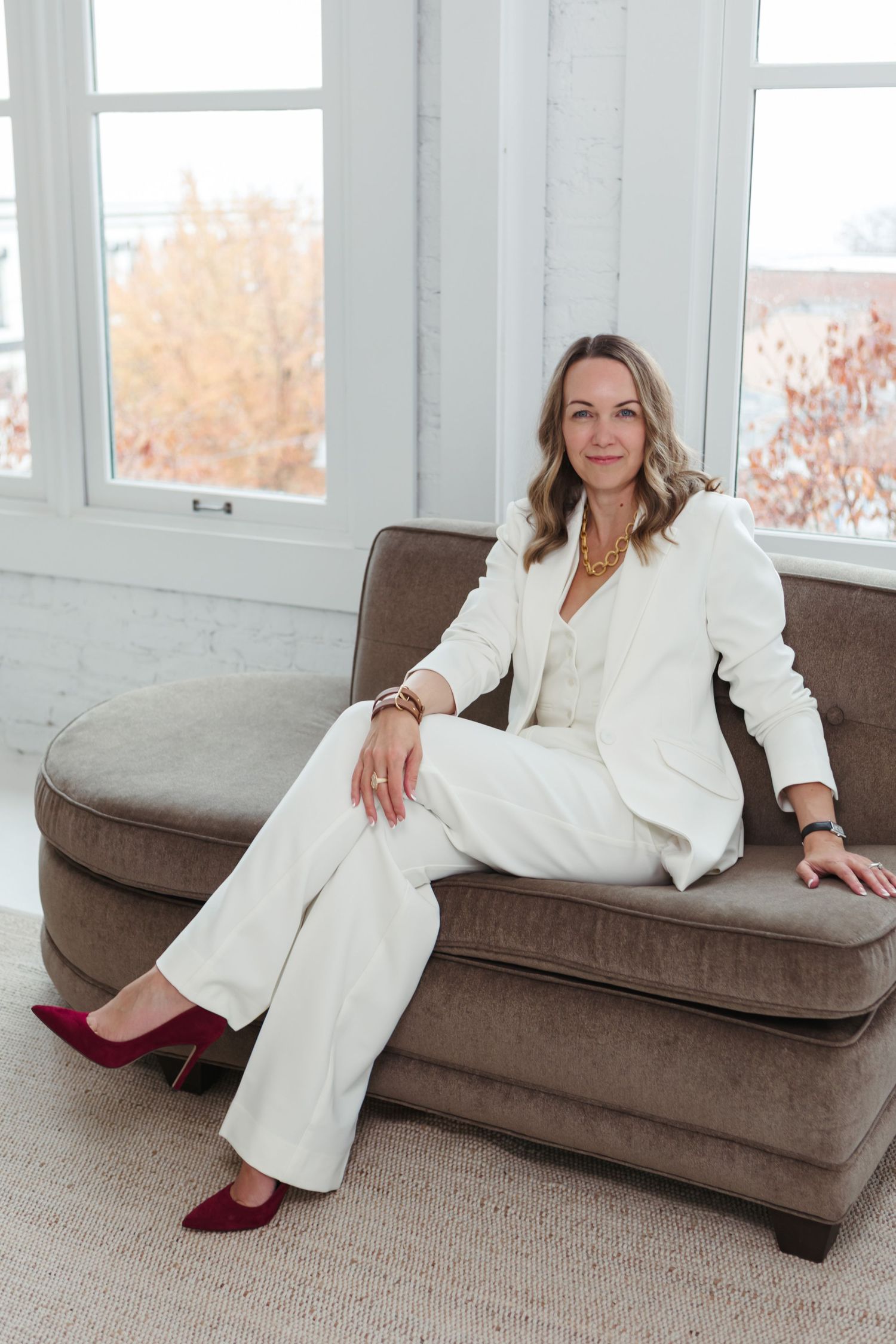 A person in a white pantsuit and burgundy shoes reclines on a brown couch near large windows in a bright room.