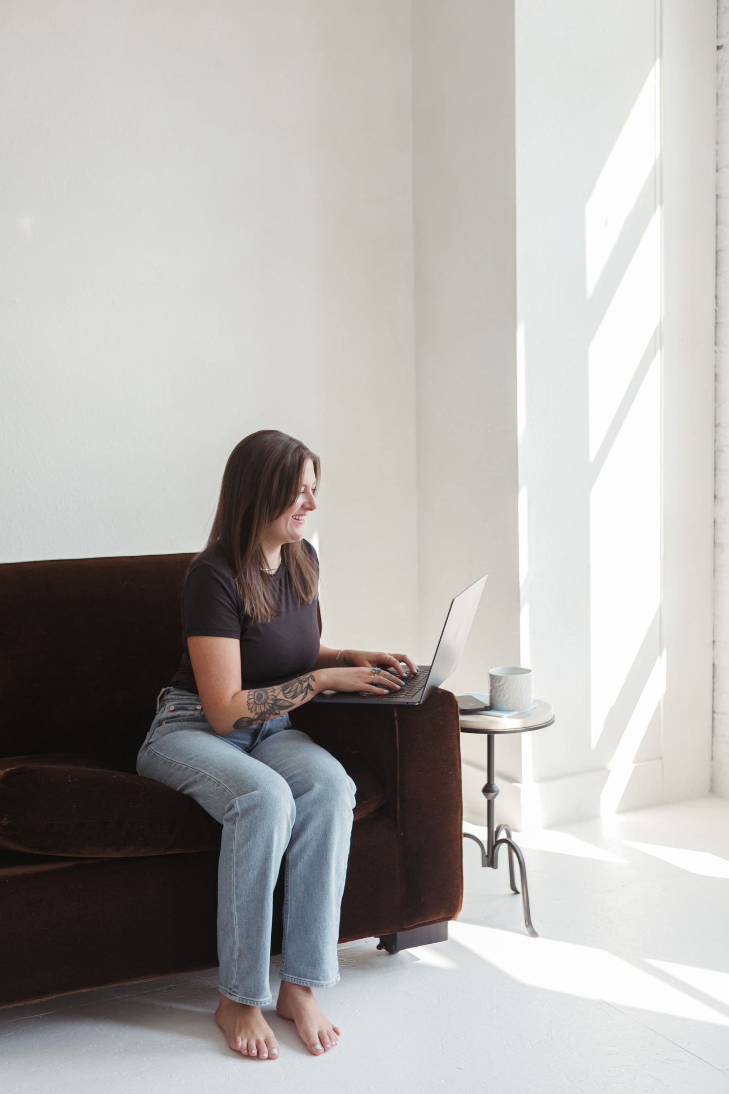 Person in casual attire sits barefoot on dark couch working on laptop in bright minimalist white room.