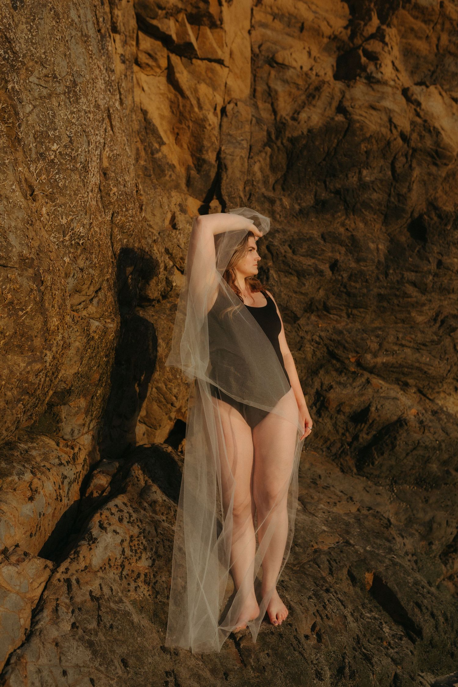 A figure in dark clothing poses against a textured rock wall with flowing sheer fabric.