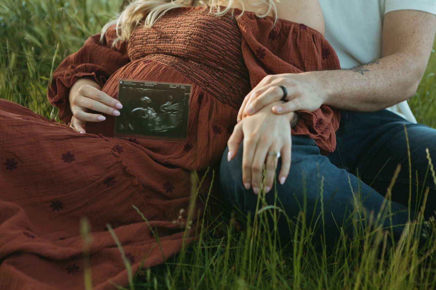 A person in a rust-colored dress sits in tall green grass while holding an ultrasound photo.