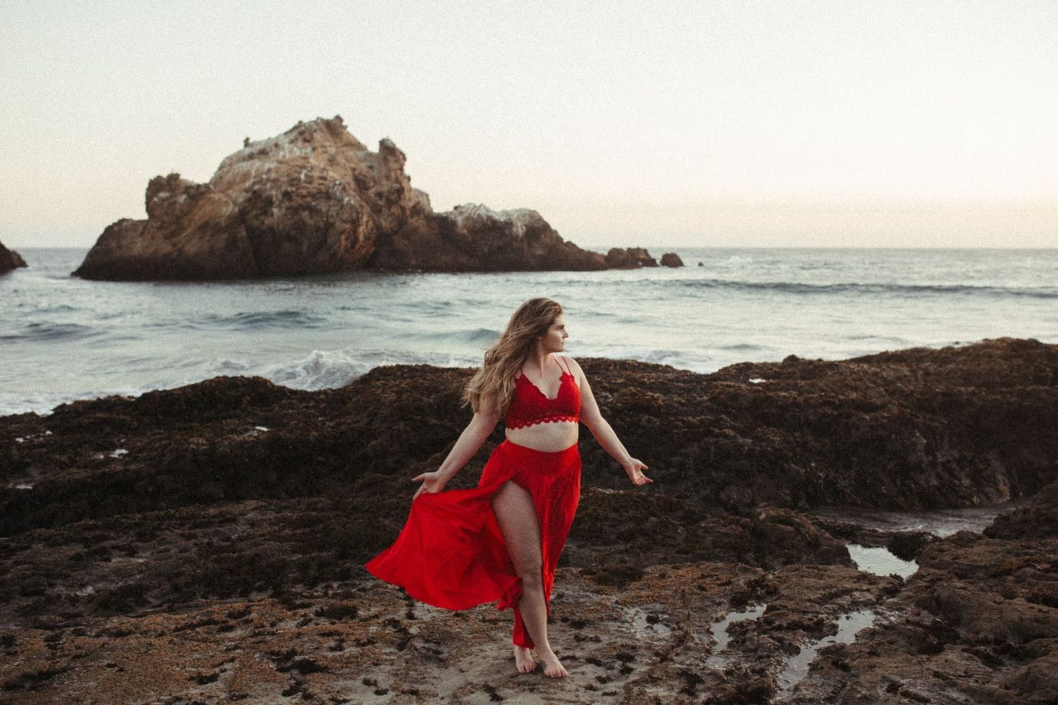 A flowing red dress catches the wind on rocky coastal cliffs at sunset with ocean waves in the background.