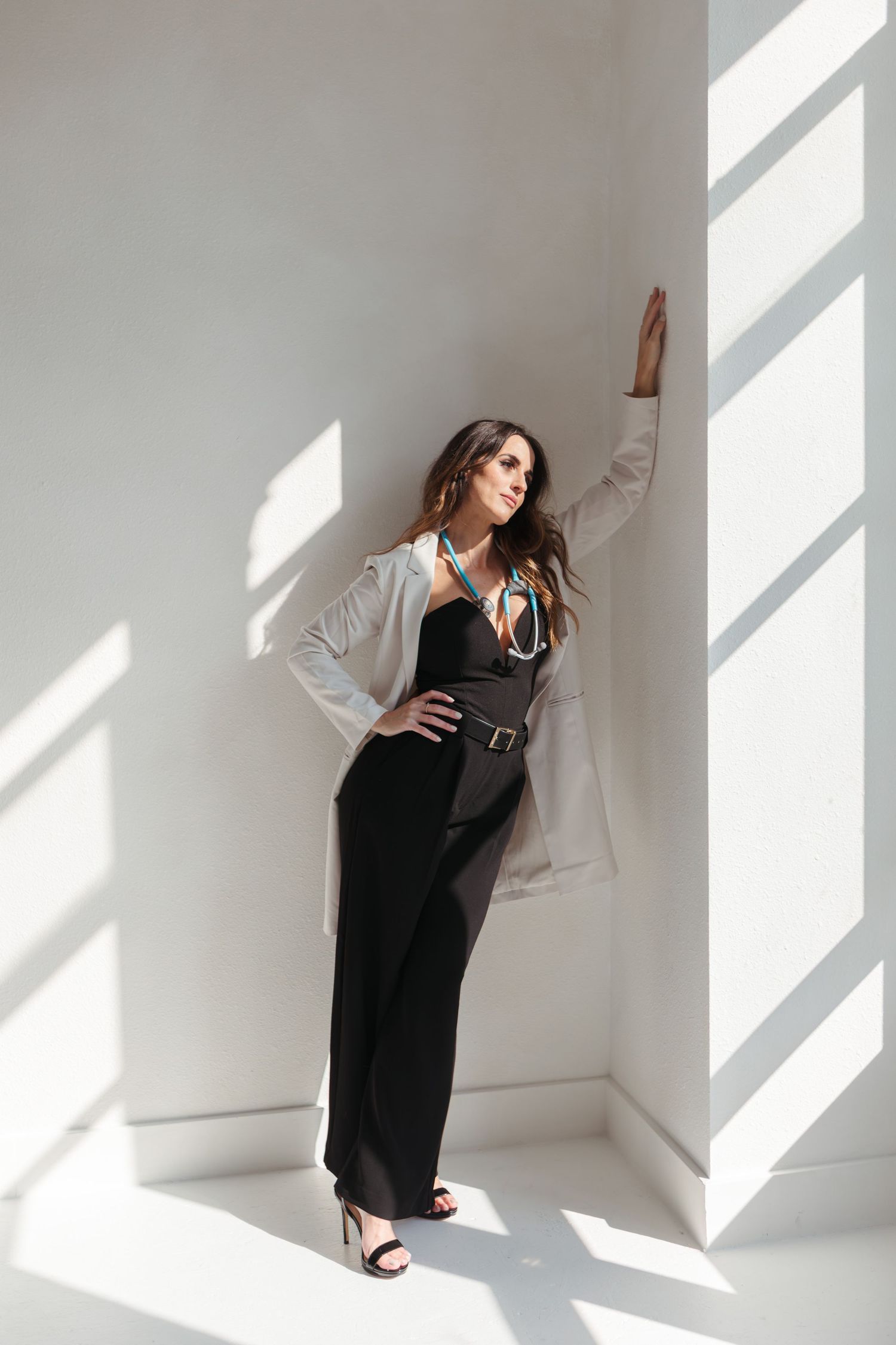 Fashion model in black jumpsuit and beige blazer poses against white wall with dramatic window lighting.