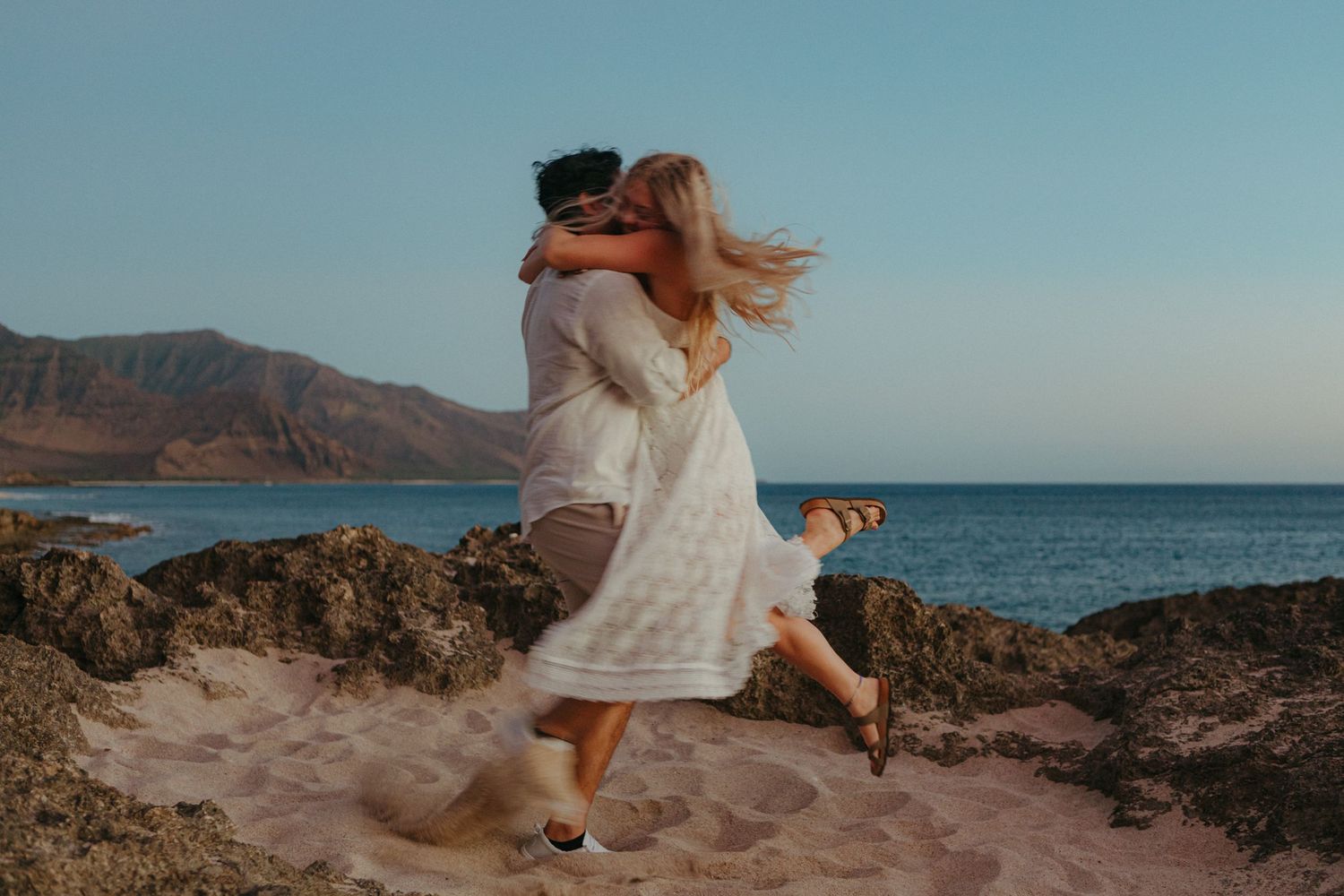 Couple embracing and kissing on a sandy beach with rocky shores and ocean views at golden hour.