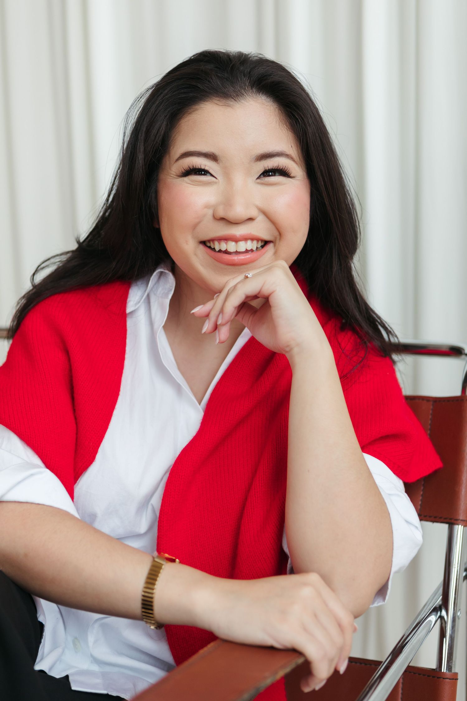 Smiling woman with long dark hair, a white shirt and red cardigan draped over her shoulders, resting her chin on her hand in a chair.