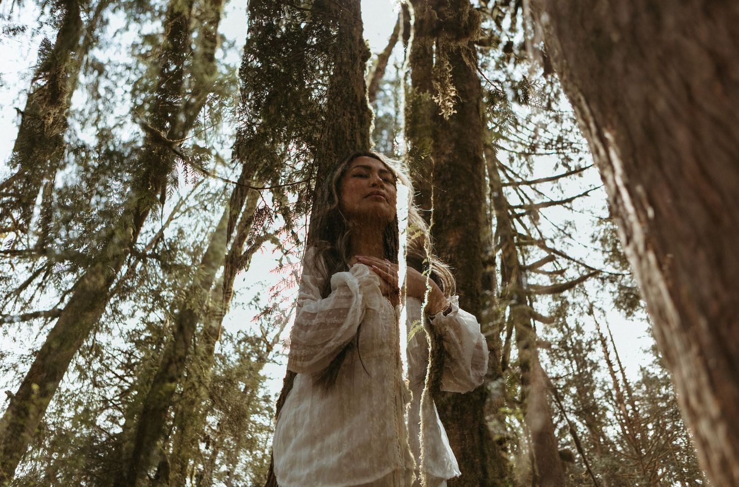 A person in a white dress stands among tall trees in a forest during a dreamy outdoor photo session.