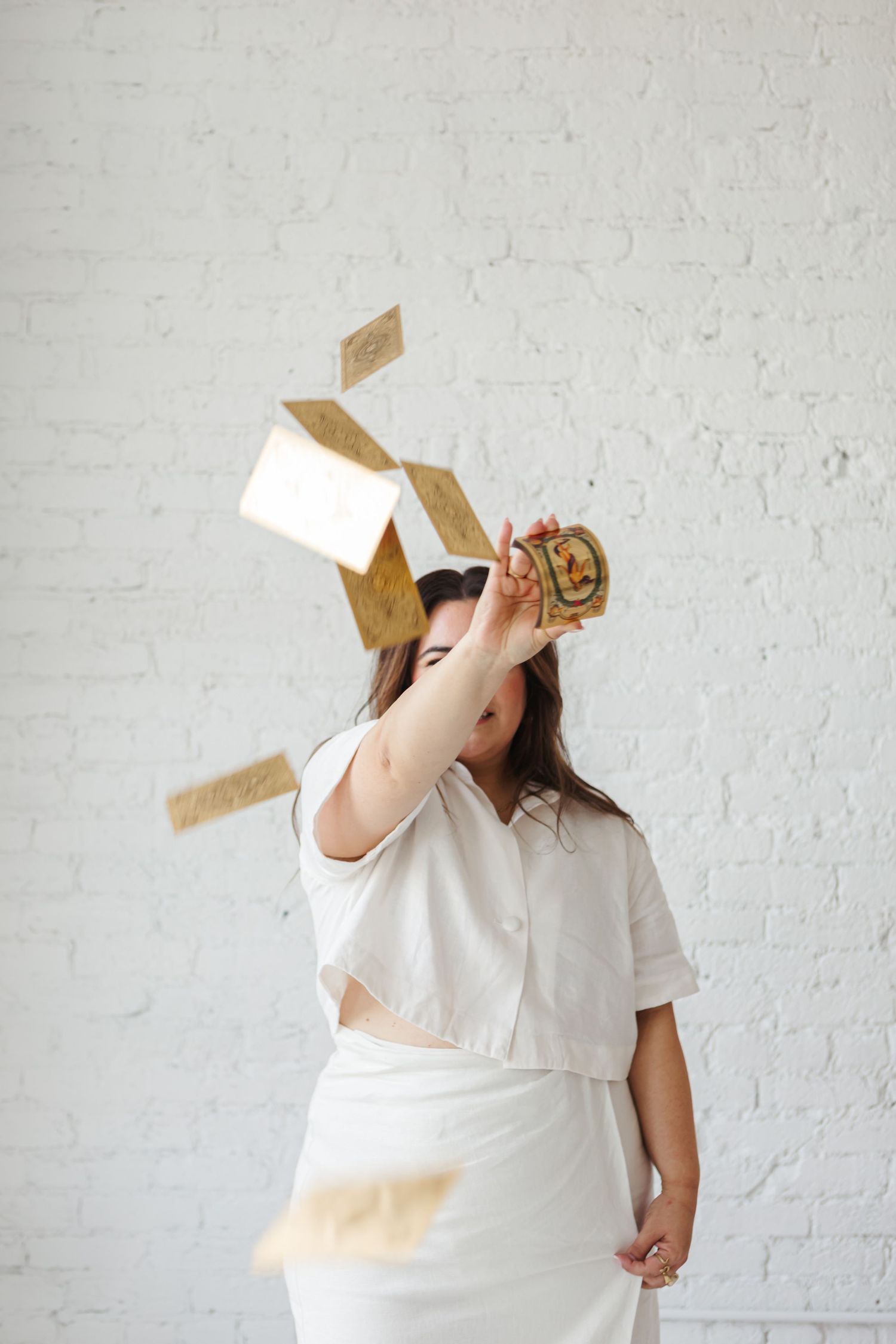 A person in white clothing playfully covers their face with brown paper pieces against a white brick wall.