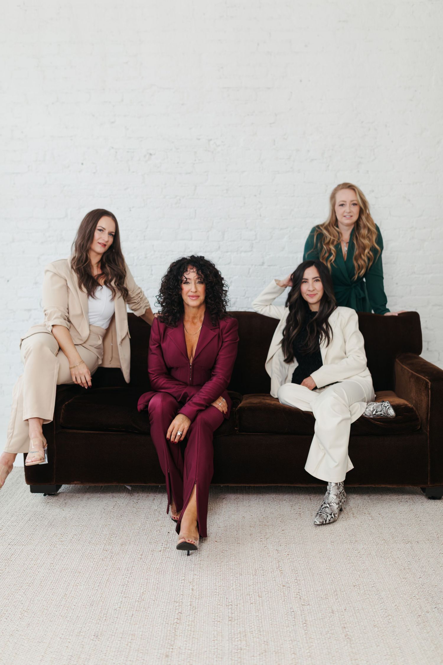 A group of women in business attire sit casually on a brown leather couch against a white wall.