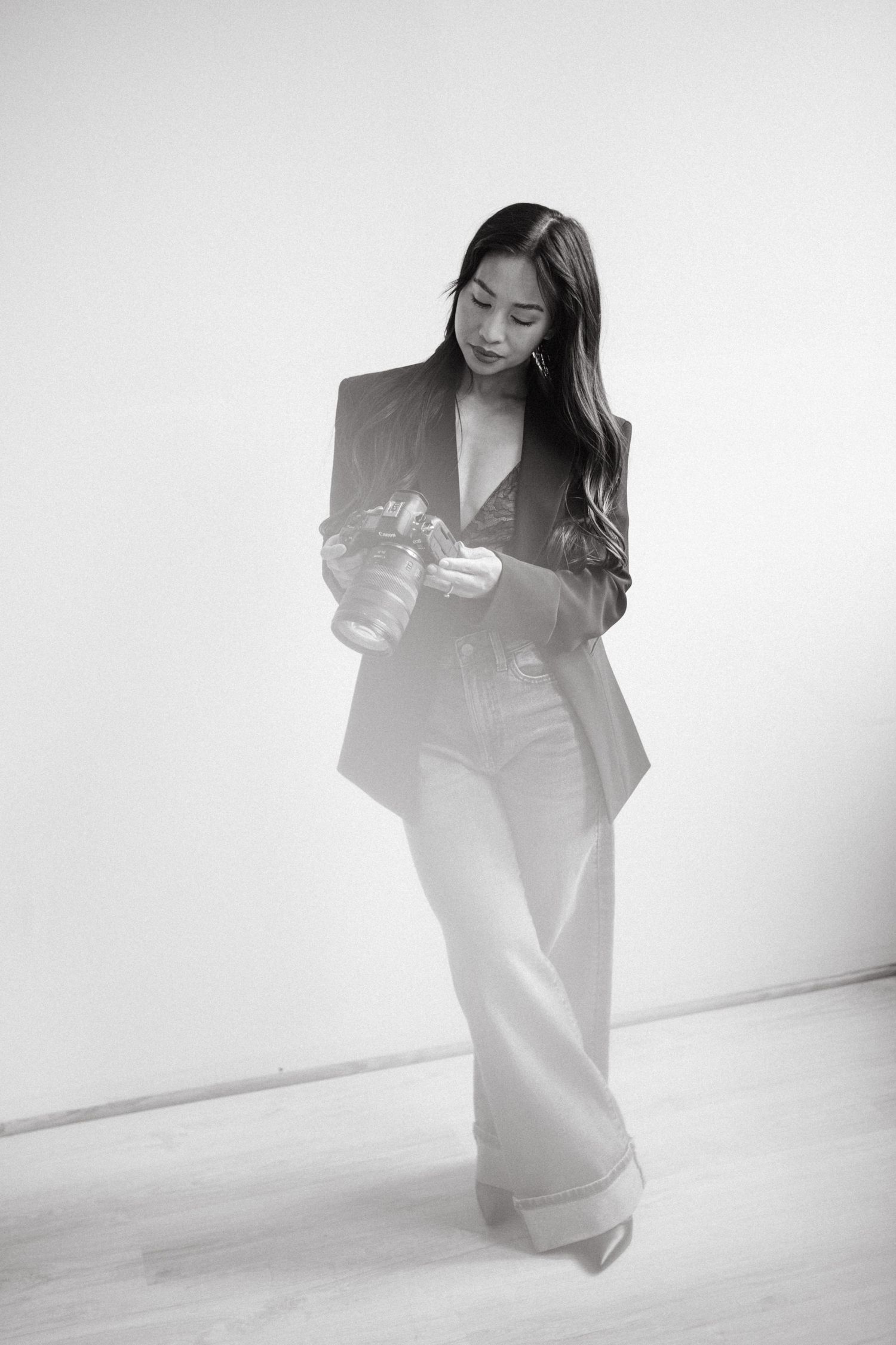 Black and white photo of a woman in a tailored blazer and wide-leg trousers standing in a bright studio, looking down at her camera.