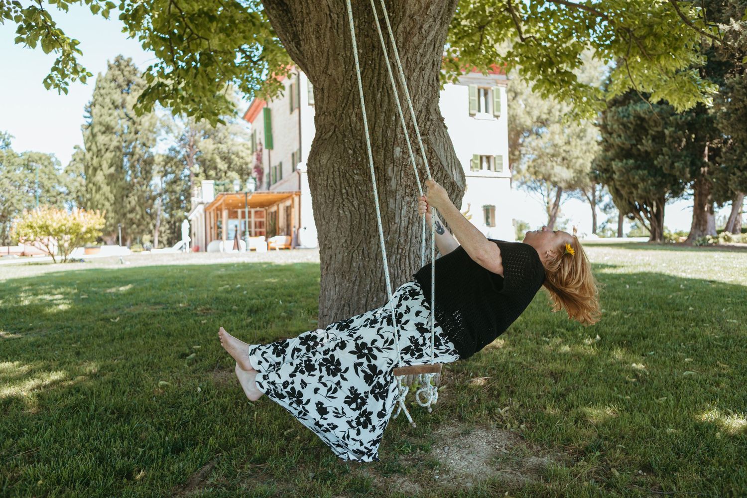 Woman in floral skirt swinging on a rope swing tied to a large tree in a sunny park.