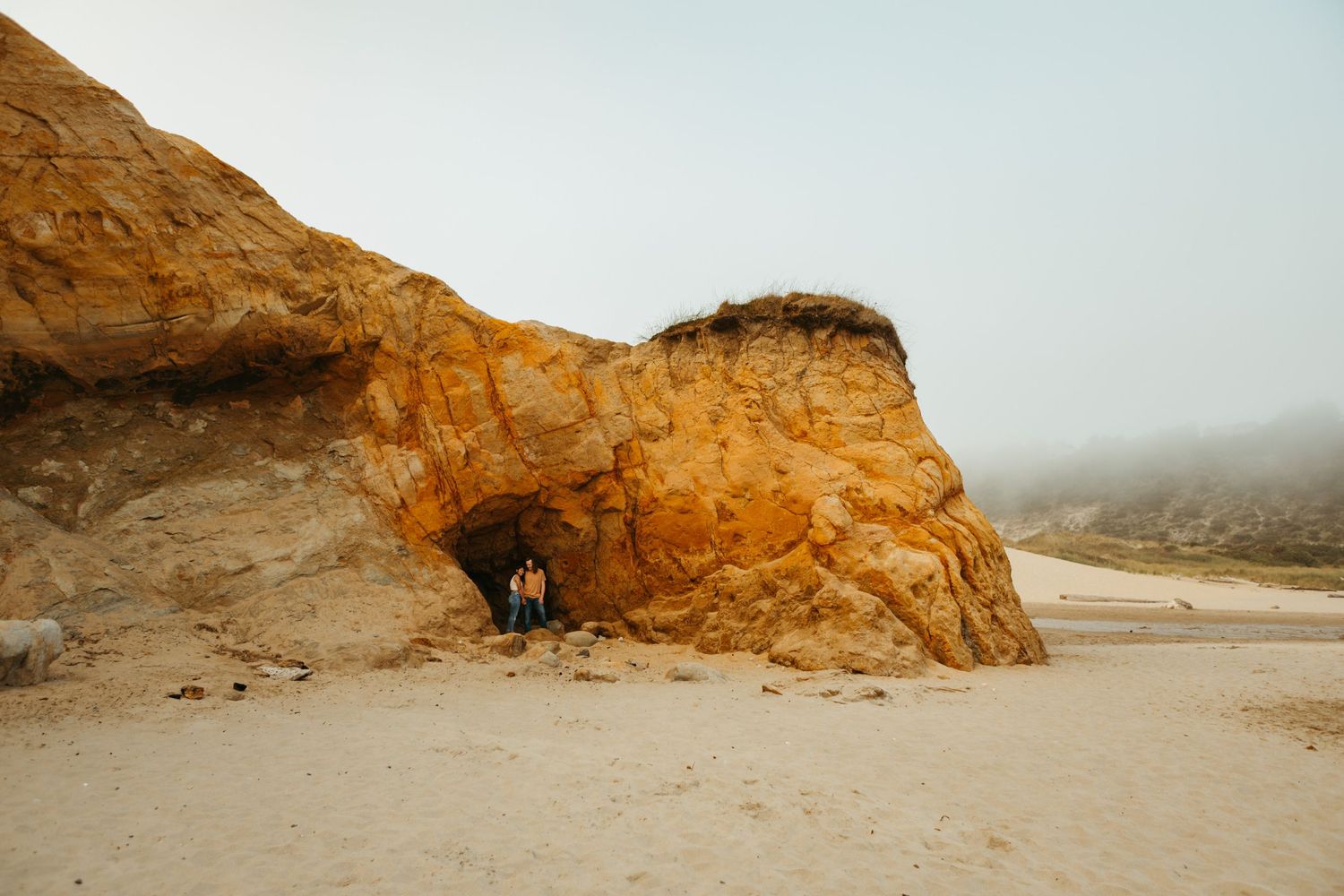 Two people stand beneath a dramatic golden sandstone sea cave arch on a misty sandy beach.