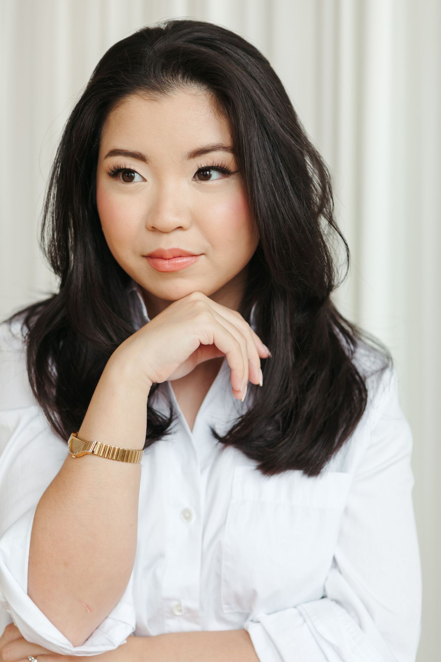 Woman with long dark wavy hair in a white button-up shirt and gold watch rests her chin on her hand against a soft curtain backdrop.